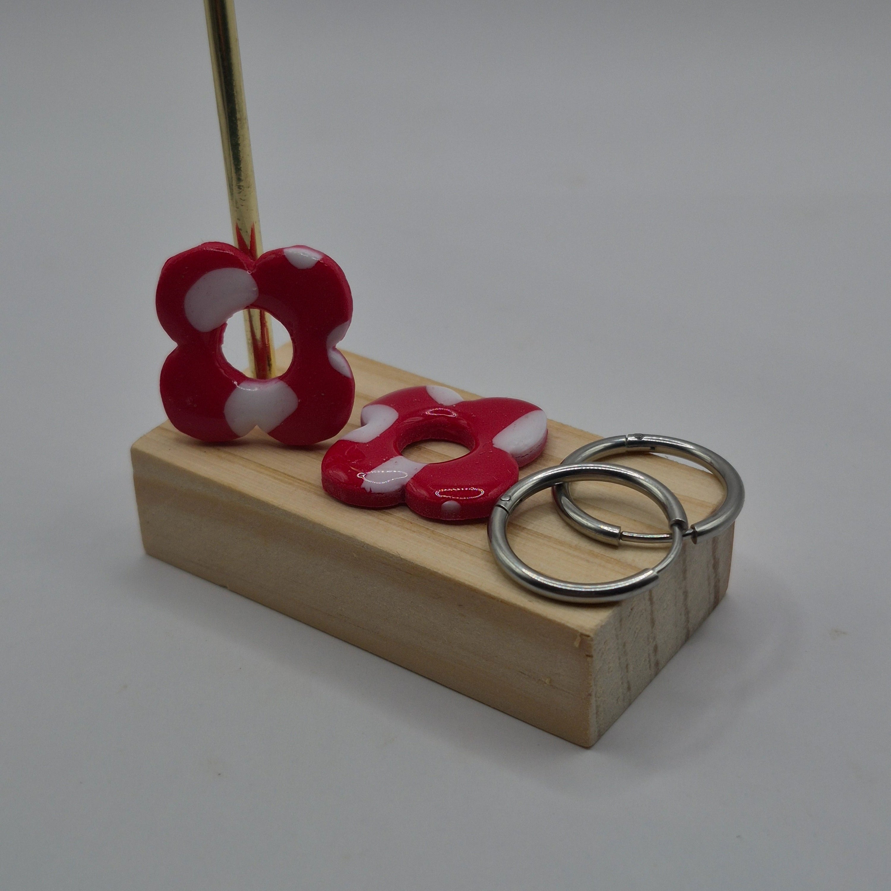 Parure Carine : pampilles interchangeables en pâte polymère rouge et blanc, en forme de fleur, présentées sur un support en bois avec deux créoles en acier inoxydable argentées, et en arrière-plan des branches de sapin, des guirlandes et des boules de Noël.