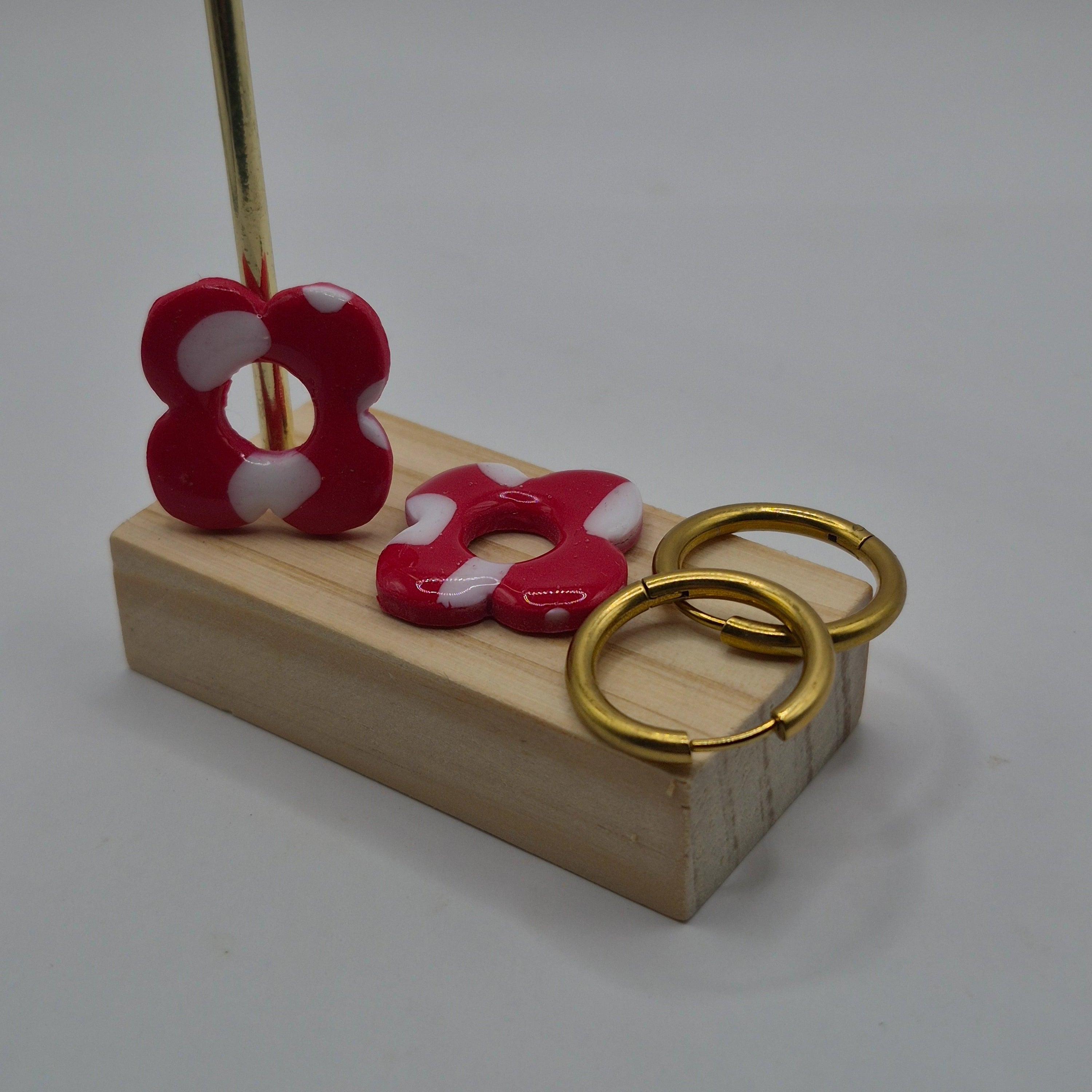 Parure Carine : pampilles interchangeables en pâte polymère rouge et blanc, en forme de fleur, présentées sur un support en bois avec deux créoles en acier inoxydable dorées, et en arrière-plan des branches de sapin, des guirlandes et des boules de Noël.