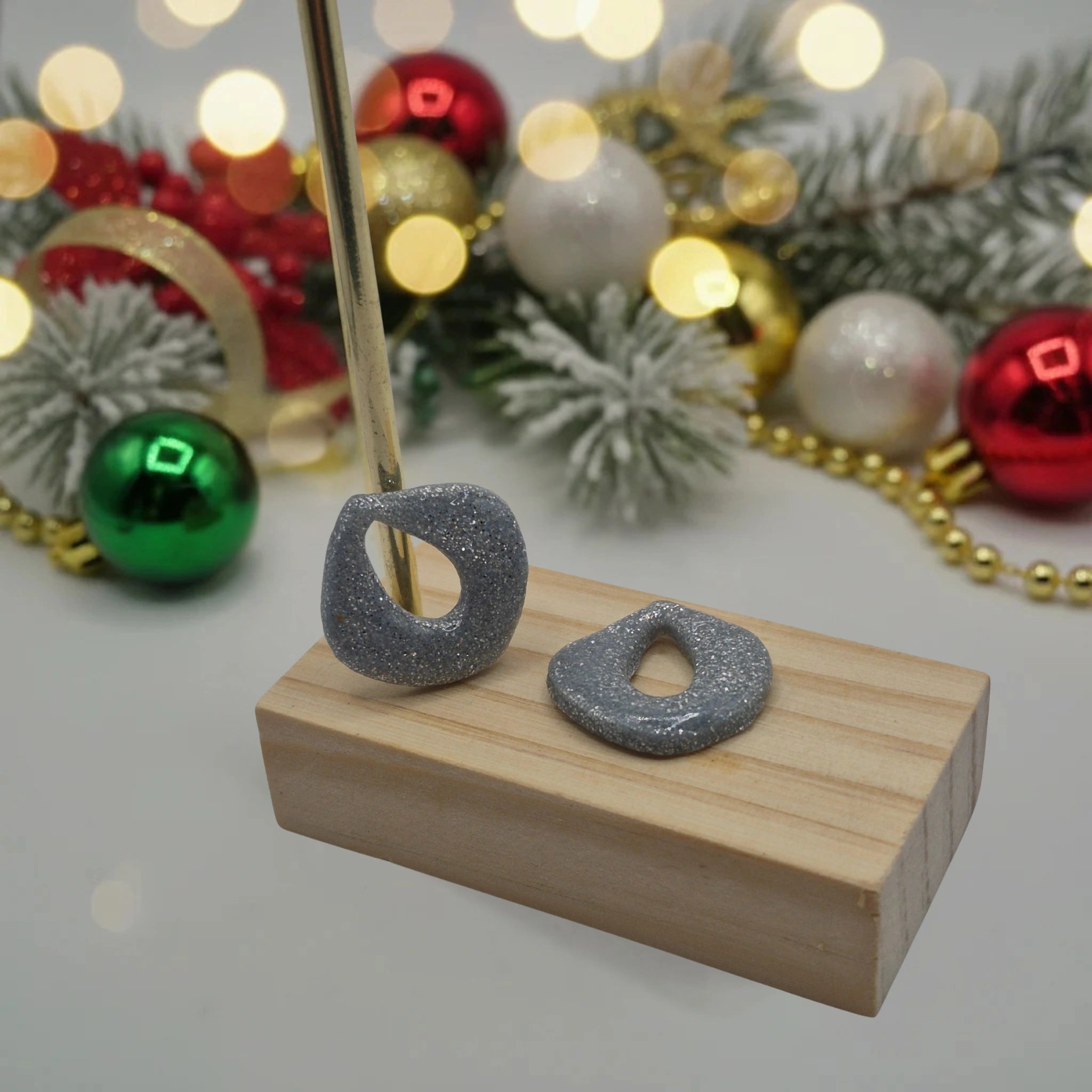 Parure Cynthia : pampilles interchangeables en pâte polymère gris argenté pailletée, en forme de goutte d'eau, présentées sur un support en bois, et en arrière-plan des branches de sapin, des guirlandes et des boules de Noël.
