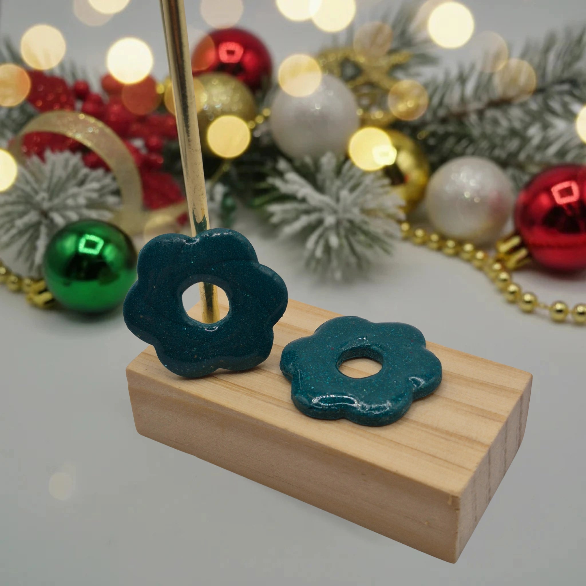 Parure Iris : pampilles interchangeables en pâte polymère vertes pailletées, en forme de fleur, présentées sur un support en bois, et en arrière-plan des branches de sapin, des guirlandes et des boules de Noël.