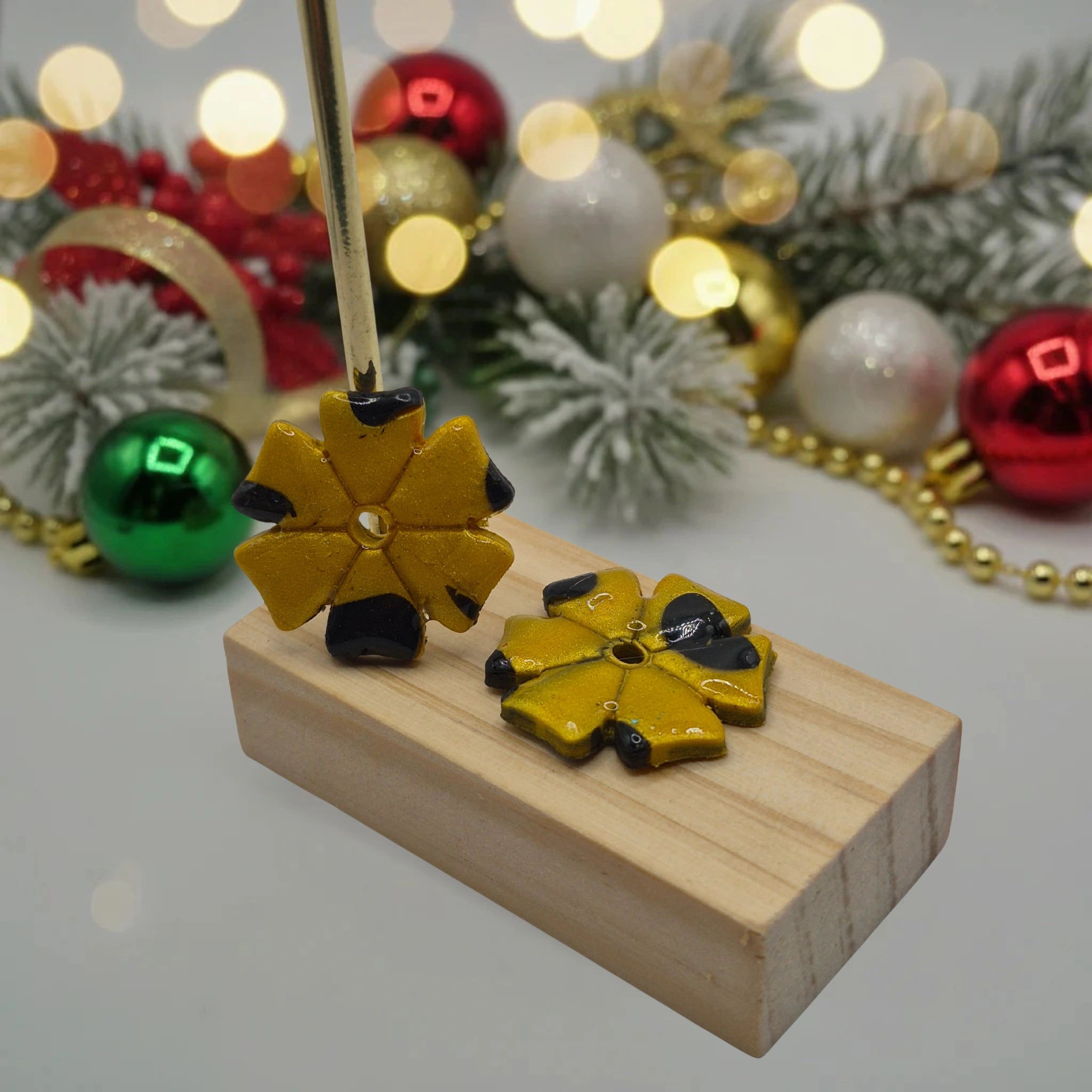 Parure Jessica : pampilles interchangeables en pâte polymère doré et noir, en forme de trèfle à 6 feuilles, présentées sur un support en bois, et en arrière-plan des branches de sapin, des guirlandes et des boules de Noël.