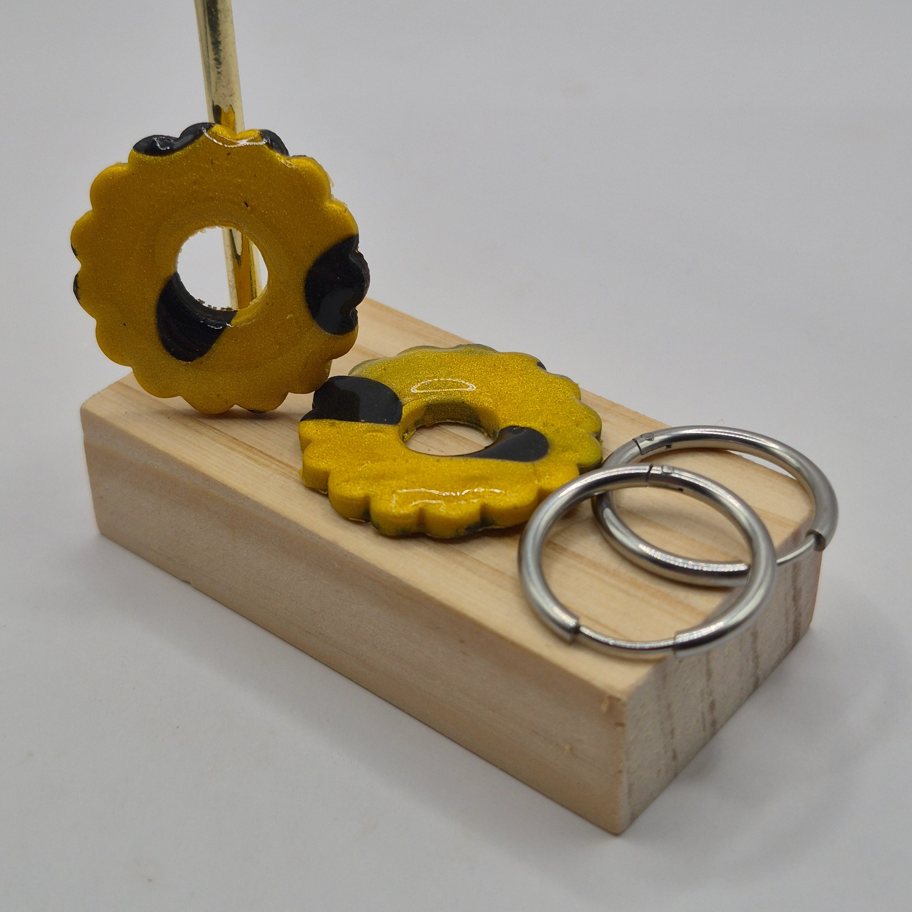 Parure Mélissa : pampilles interchangeables en pâte polymère noire et dorée pailletée, en forme de fleur, présentées sur un support en bois, avec 2 créoles en acier inoxydable argentées.