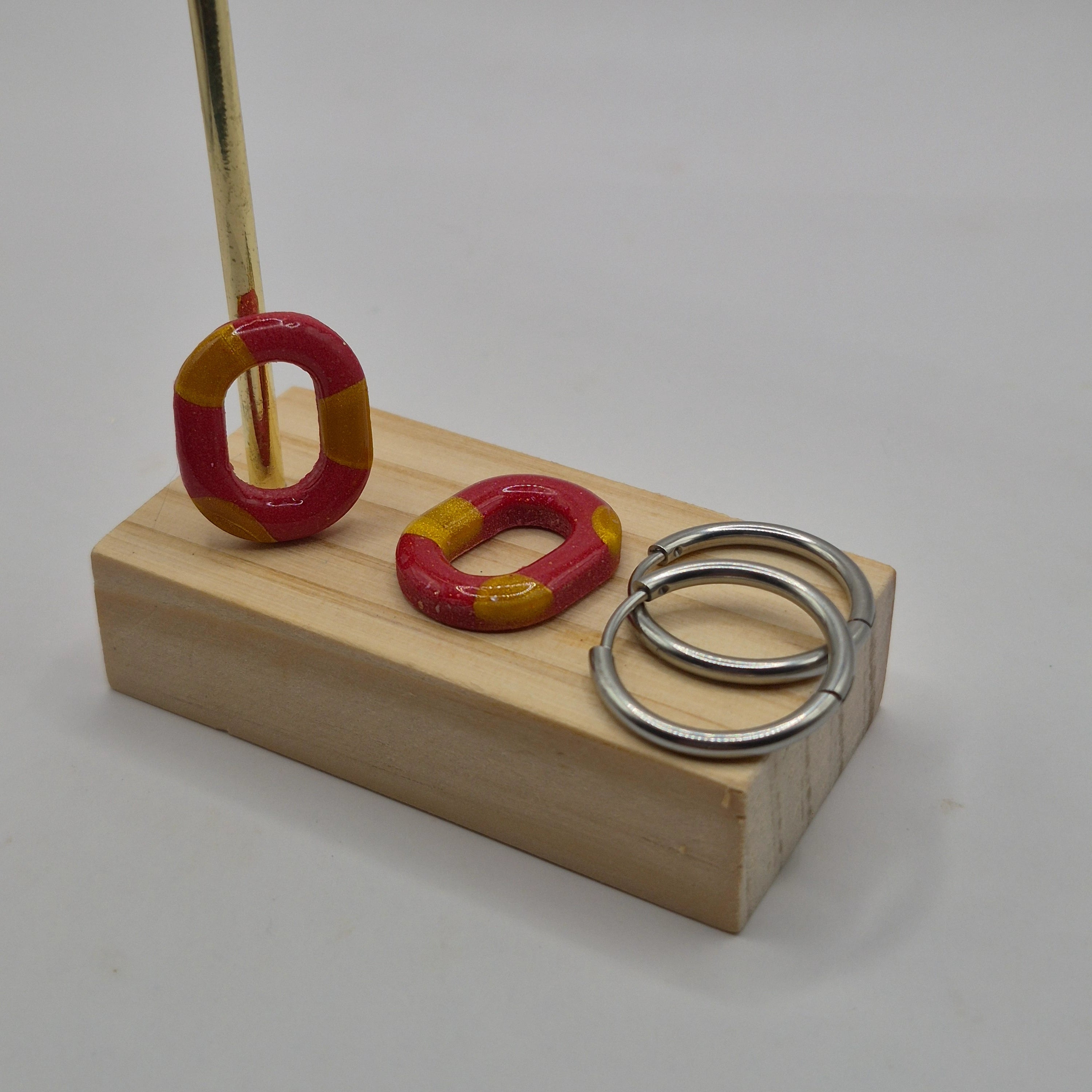 Parure Lily : pampilles interchangeables en pâte polymère rouge et doré en forme de ovale présentées sur un support en bois avec deux créoles en acier inoxydable argentées