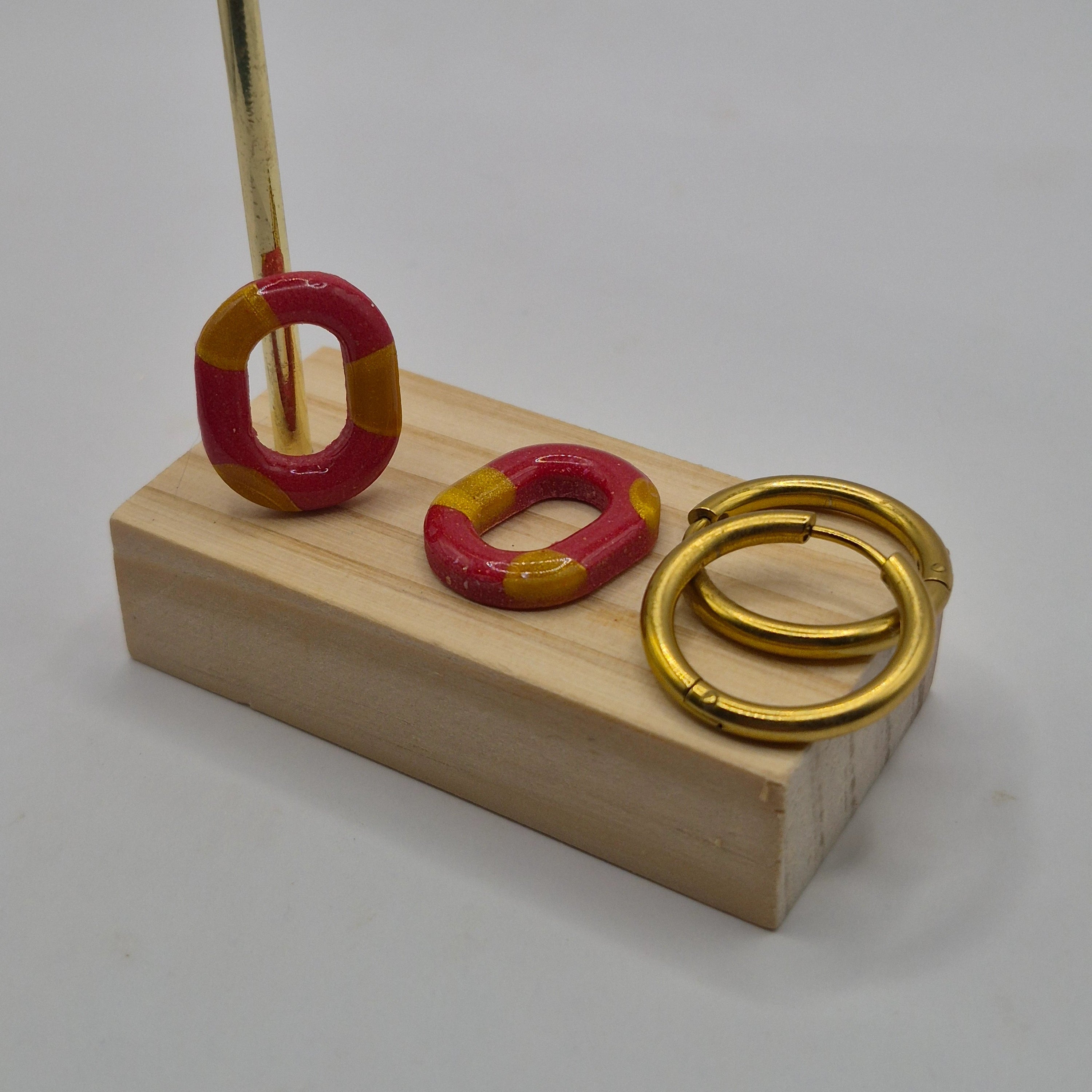 Parure Lily : pampilles interchangeables en pâte polymère rouge et doré en forme de ovale présentées sur un support en bois avec deux créoles en acier inoxydable dorées.
