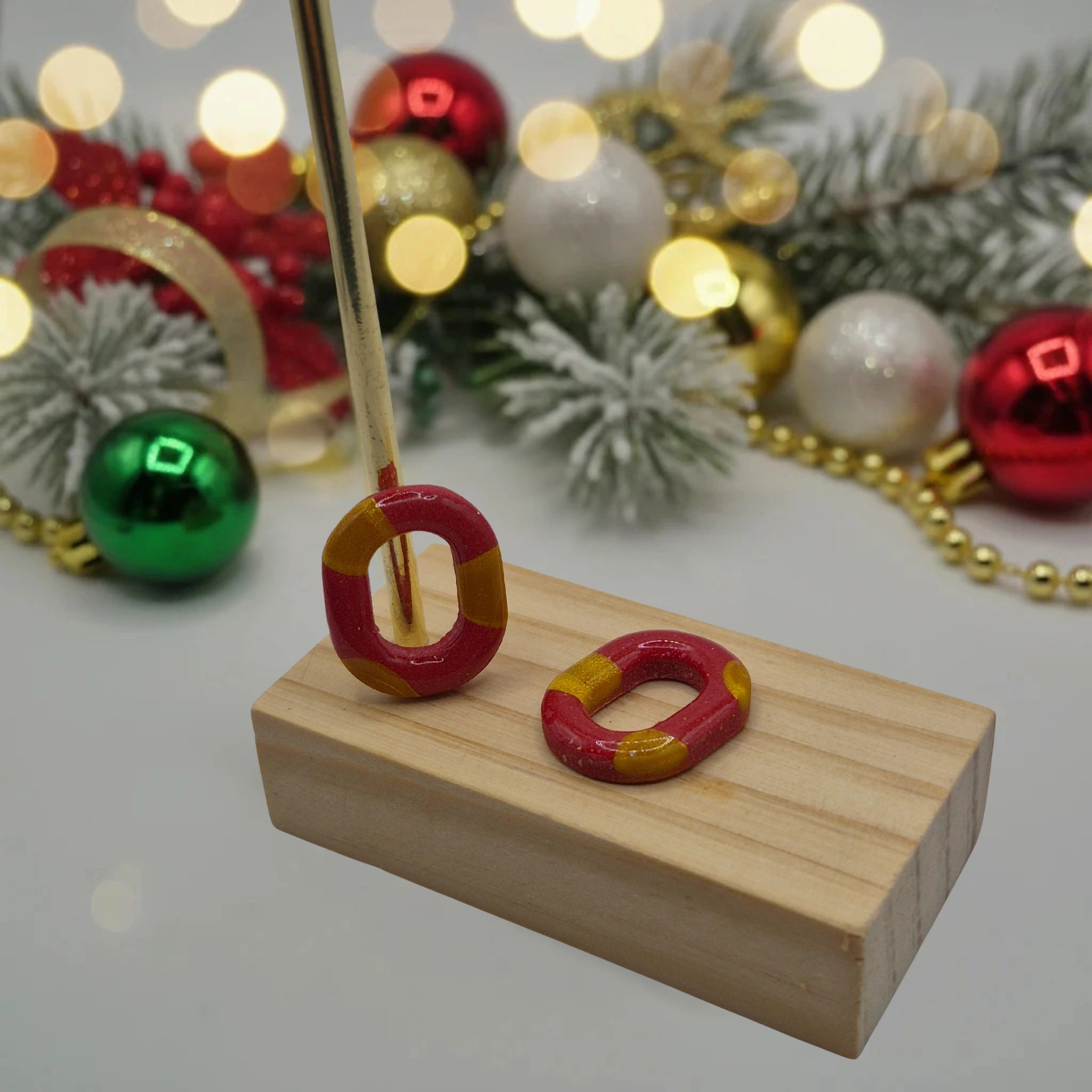 Parure Lily : pampilles interchangeables en pâte polymère rouge et blanc, en forme ovale, présentées sur un support en bois, et en arrière-plan des branches de sapin, des guirlandes et des boules de Noël.