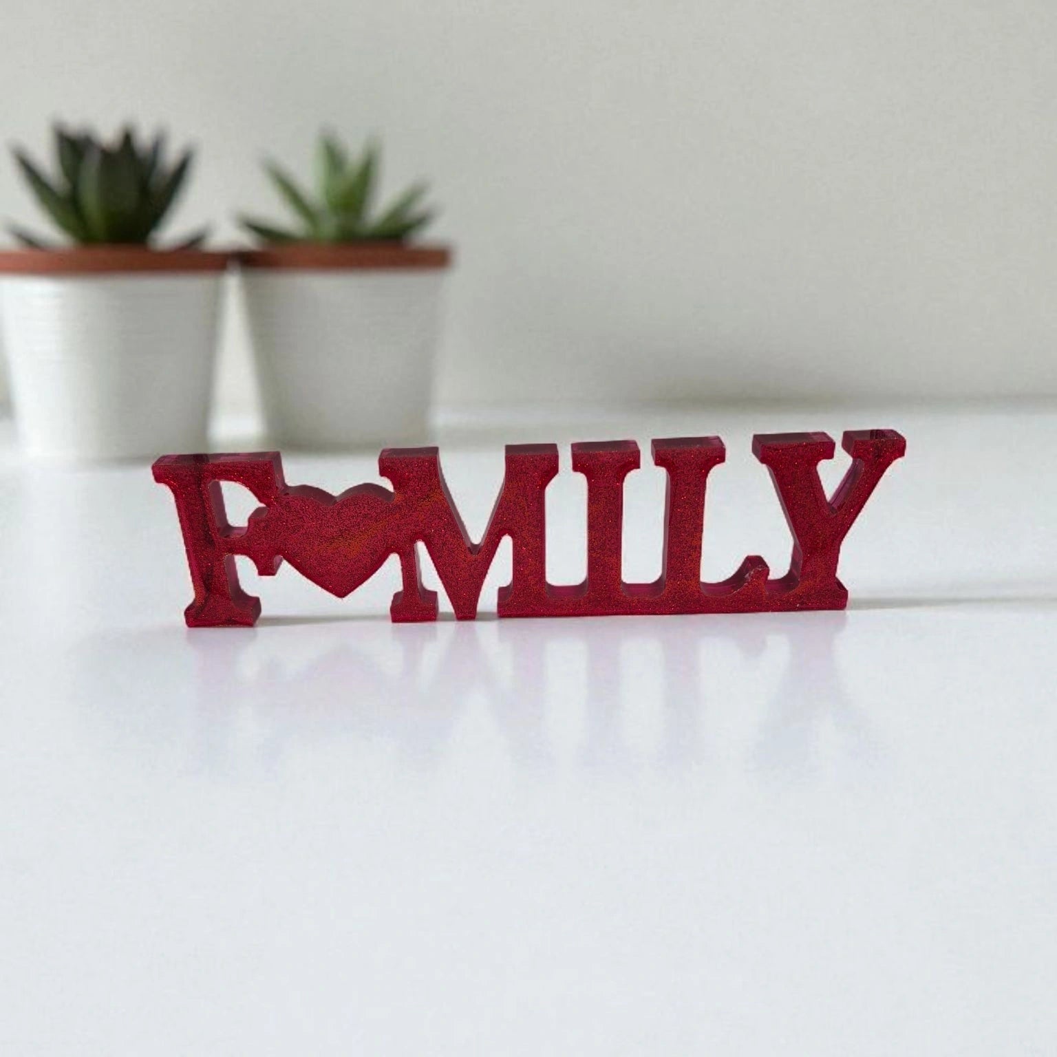 Décoration intérieure Family en résine pailletée rouge posée sur une surface plane blanche devant deux plantes.