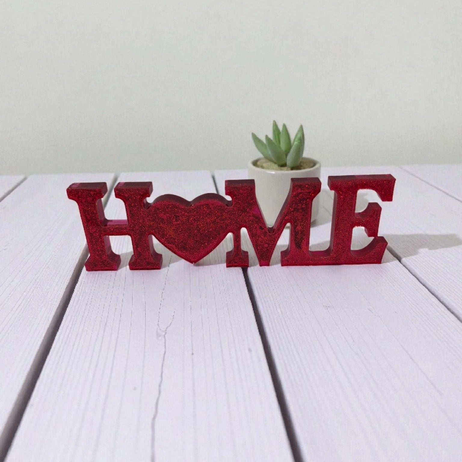 Décoration en résine époxy rouge pailletée représentant le mot « Home » avec un cœur à la place du O, posée sur une table en bois blanc devant une petite plante.