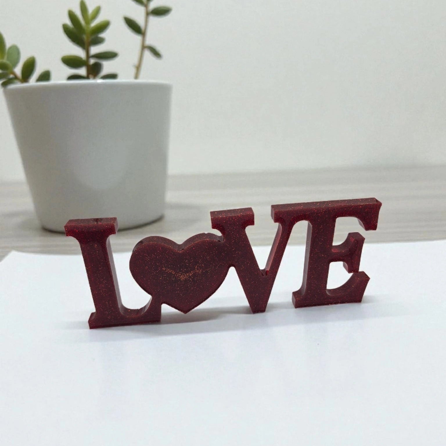 Décoration intérieure avec inscription Love en résine pailletée de couleur rouge, posée sur une feuille blanche devant plante verte