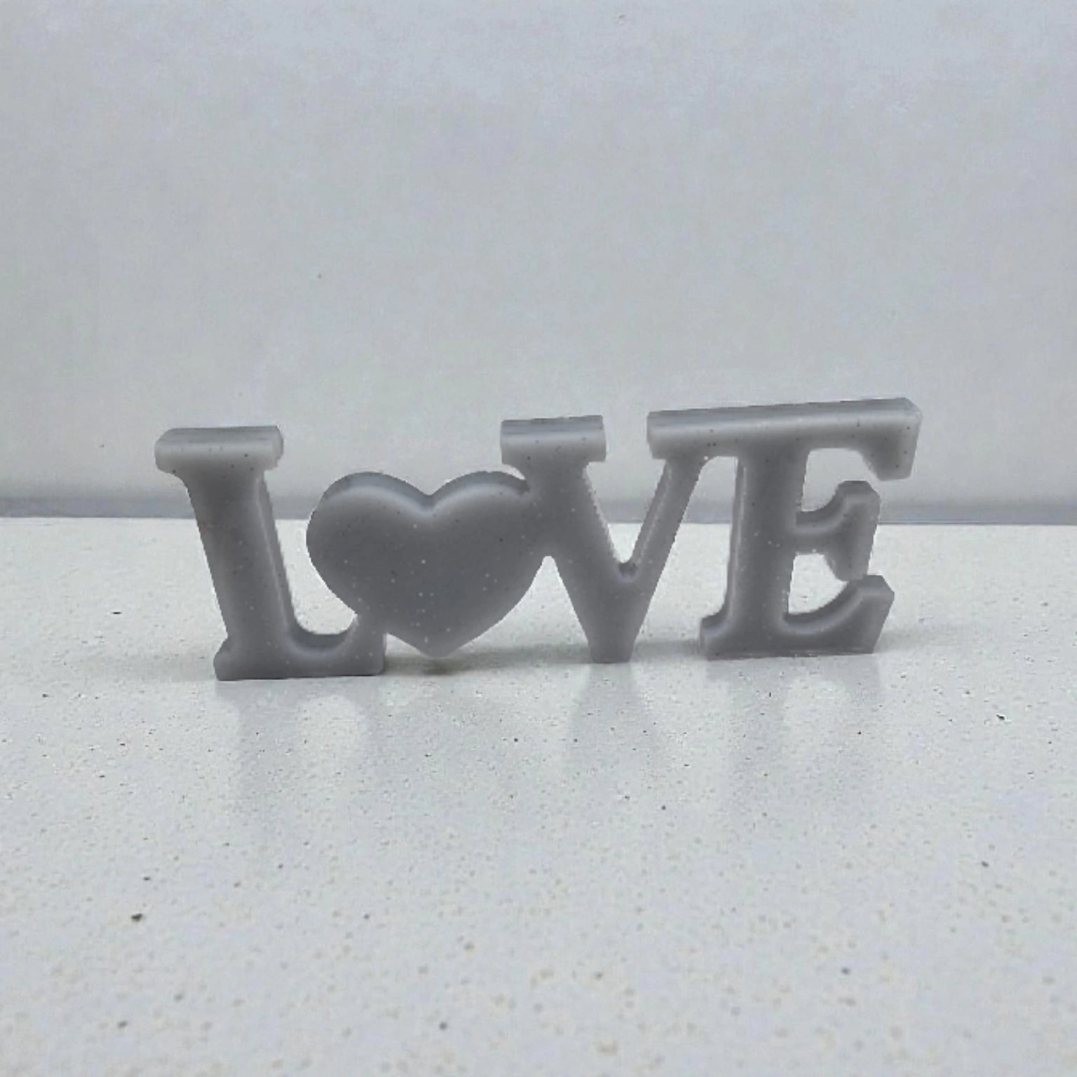 Décoration en résine époxy argent pailletée représentant le mot « Home » avec un cœur à la place du O, posée sur une surface plane blanche.