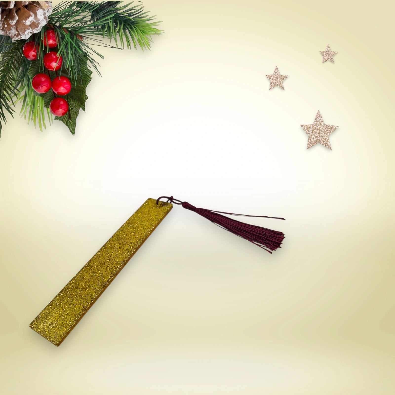 Marque-page artisanal en résine époxy dorée pailletée avec pompon bordeaux, présenté sur un fond décoré de branches de sapin et d’étoiles.