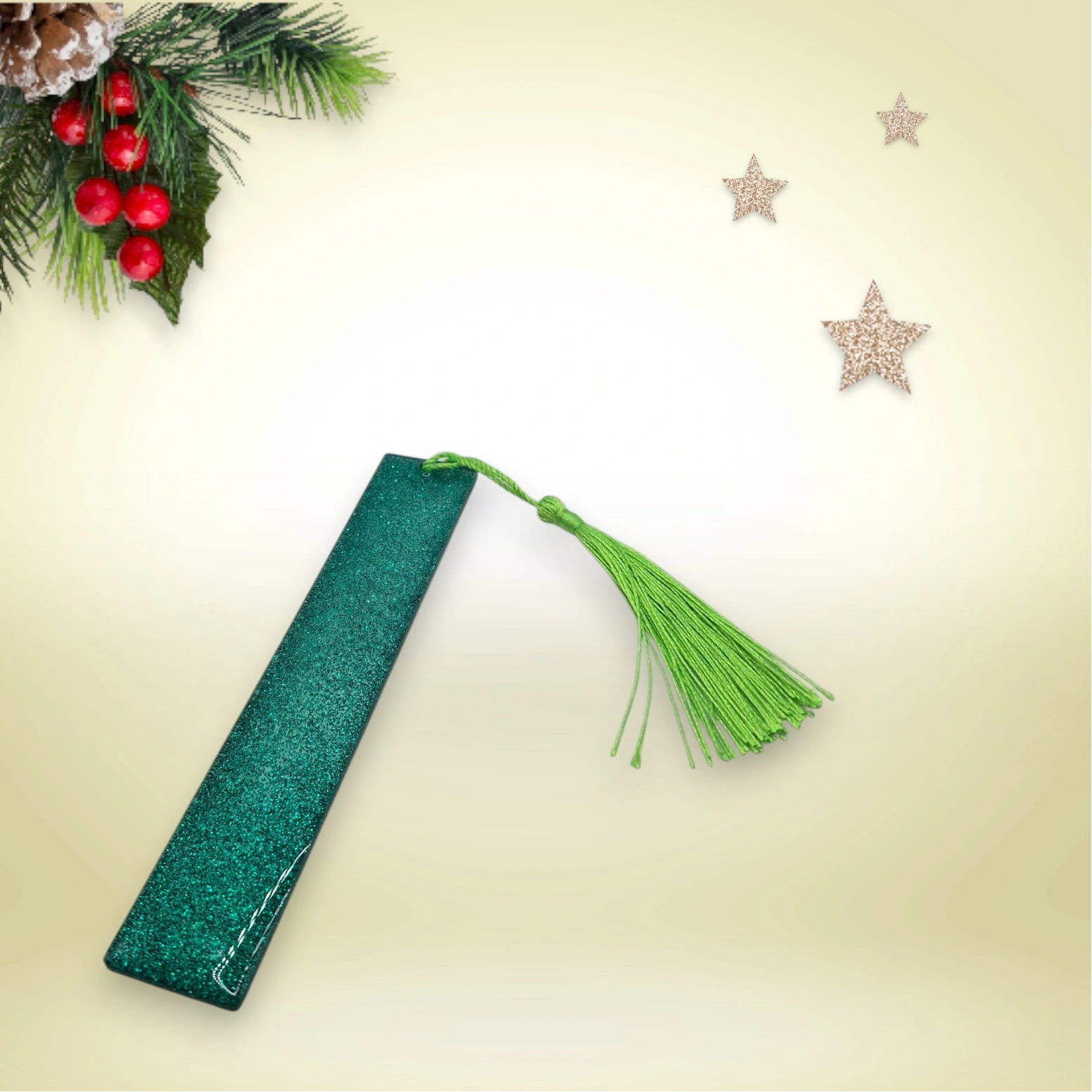 Marque-page artisanal en résine époxy verte pailletée avec pompon bordeaux, présenté sur un fond décoré de branches de sapin et d’étoiles.