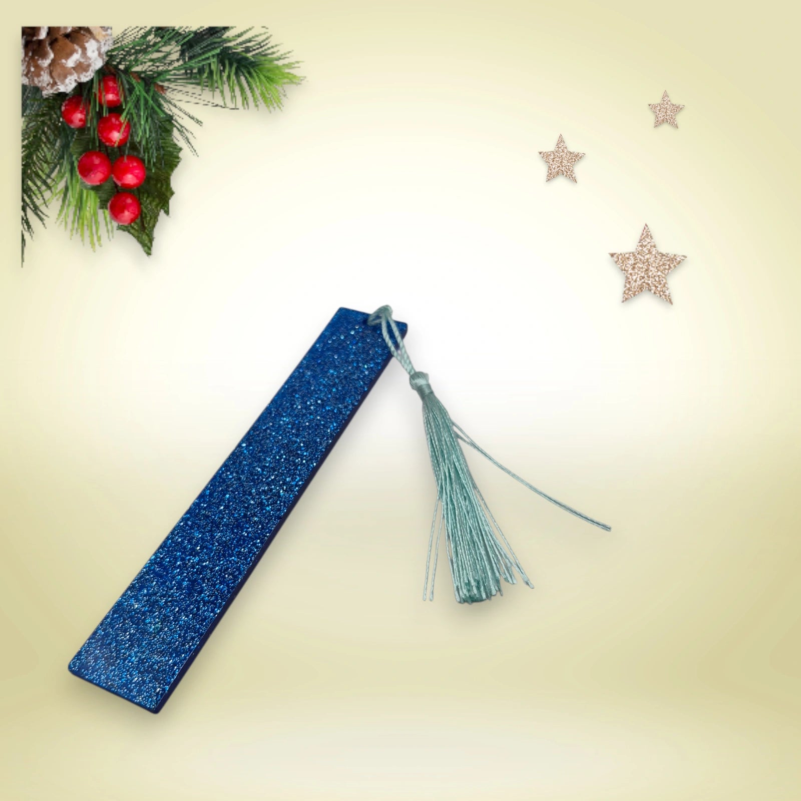 Marque-page artisanal en résine époxy bleue pailletée avec pompon bleu, présenté sur un fond décoré de branches de sapin et d’étoiles.