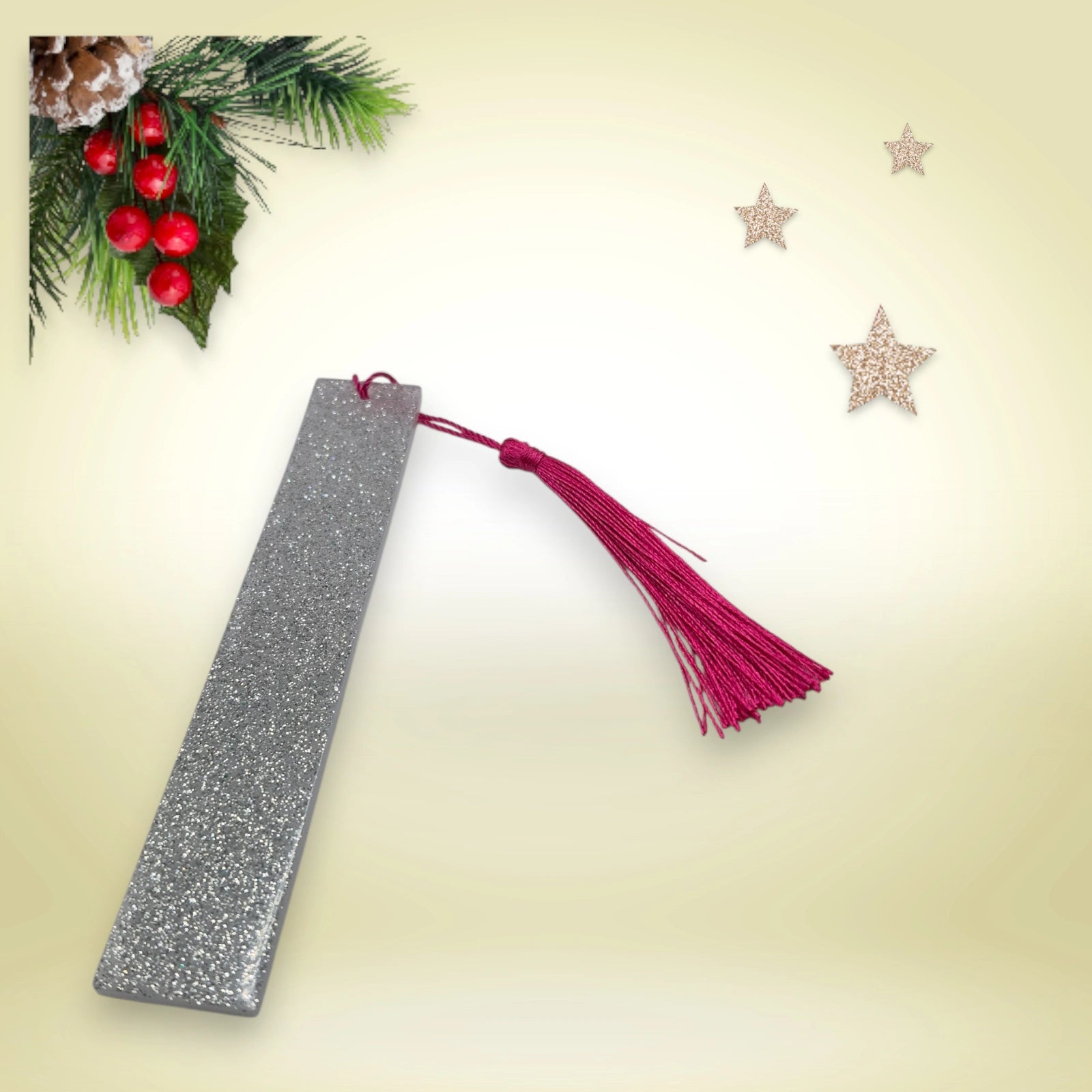 Marque-page artisanal en résine époxy argent pailletée avec pompon fuchsia, présenté sur un fond décoré de branches de sapin et d’étoiles.
