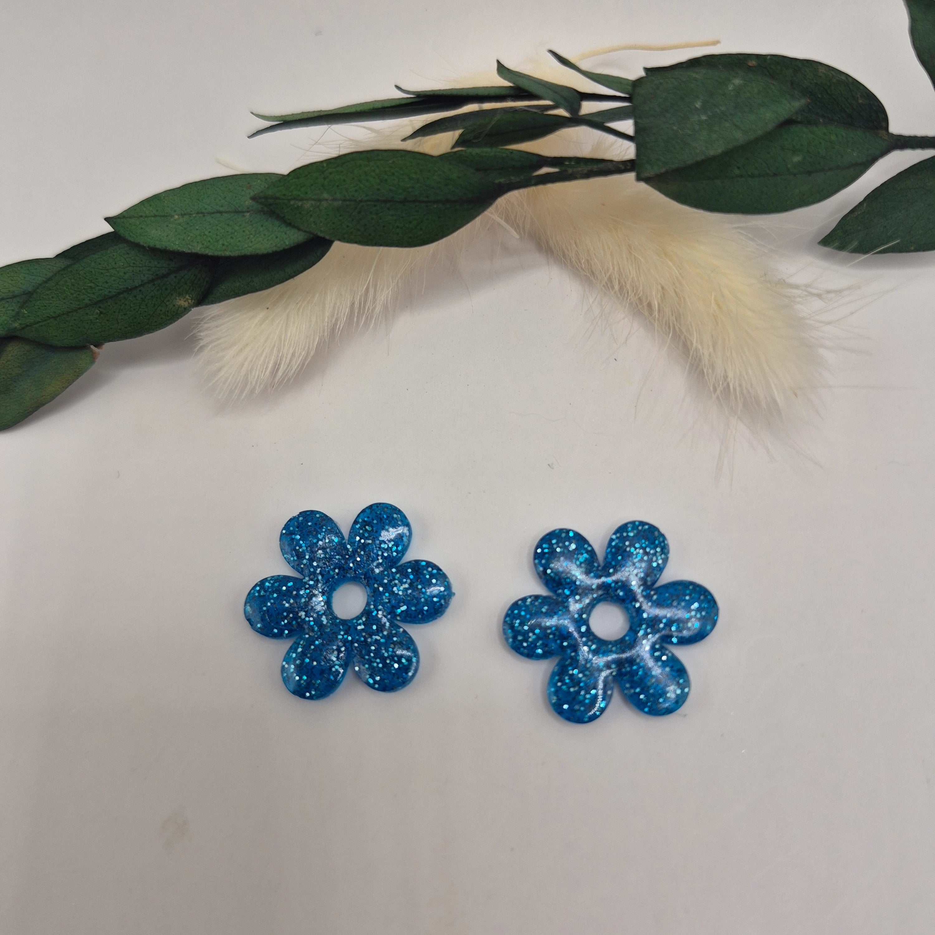 Boucles d’oreilles pampilles bleues Marjorie en résine époxy pailletée posées à plat sur fond neutre avec branche de pampa et feuilles
