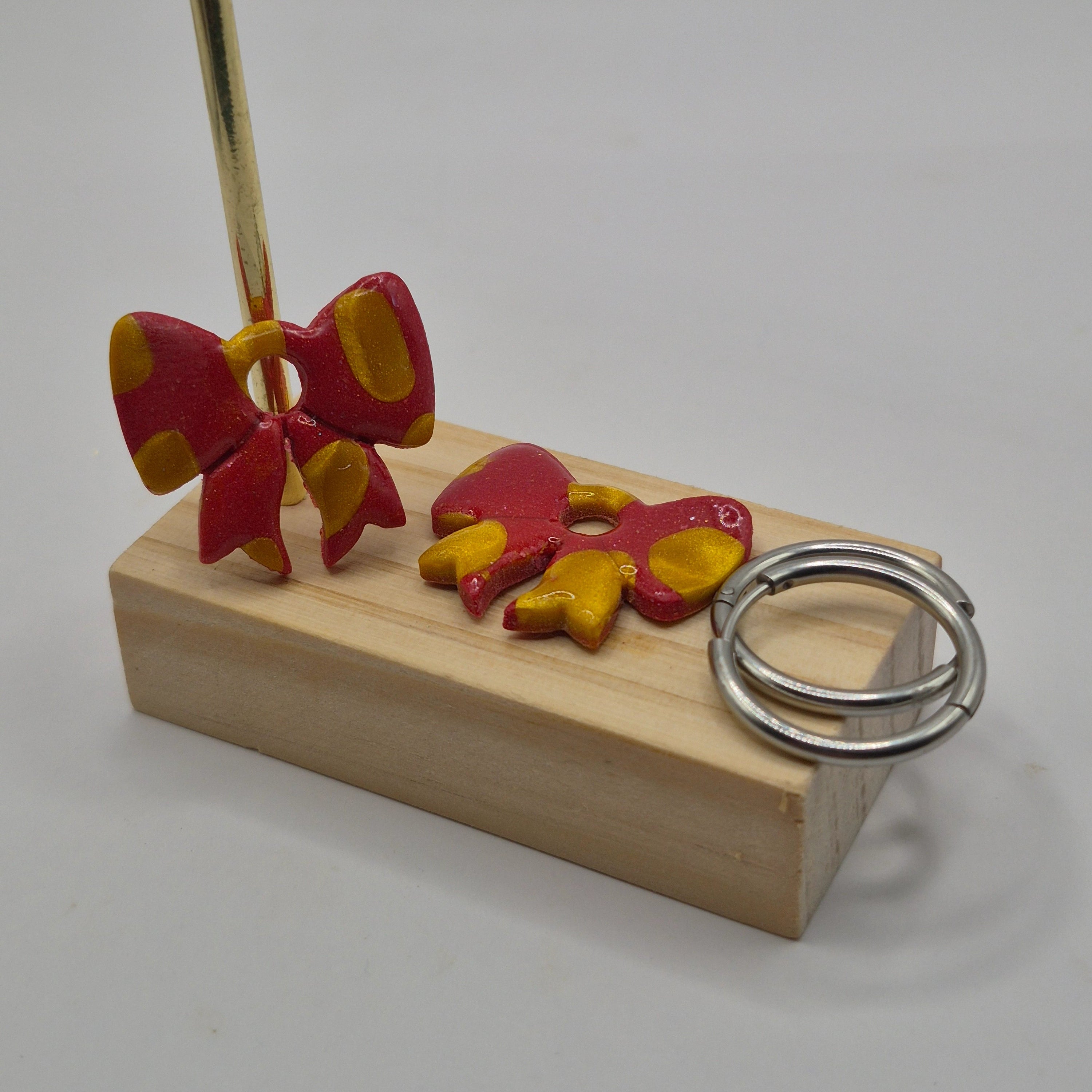 Parure Sophie : pampilles interchangeables en pâte polymère rouge et dorée pailletée, en forme de nœud, présentées sur un support en bois, avec 2 créoles argentées en acier inoxydable.