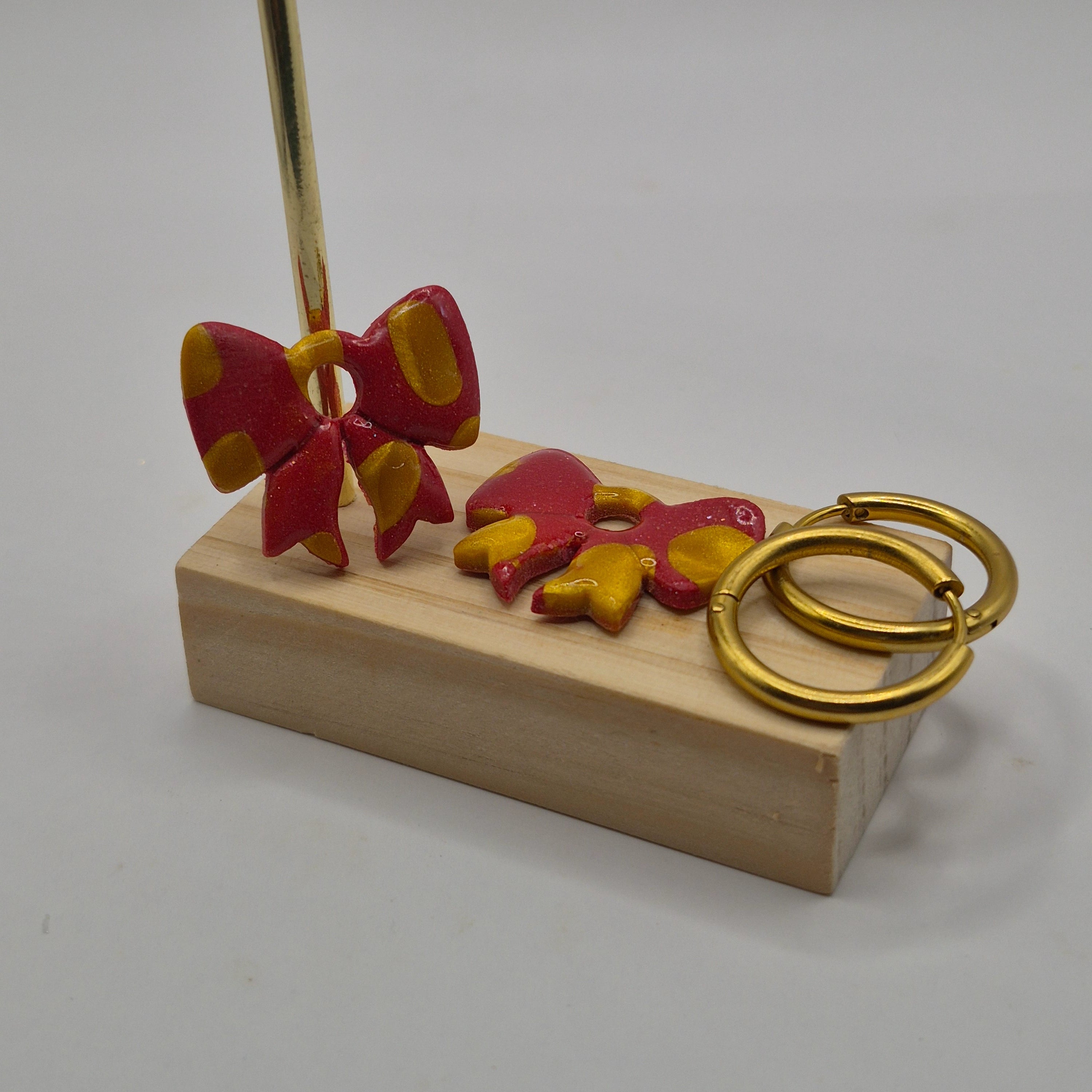 Parure Sophie : pampilles interchangeables en pâte polymère rouge et dorée pailletée, en forme de nœud, présentées sur un support en bois, avec 2 créoles dorées en acier inoxydable.