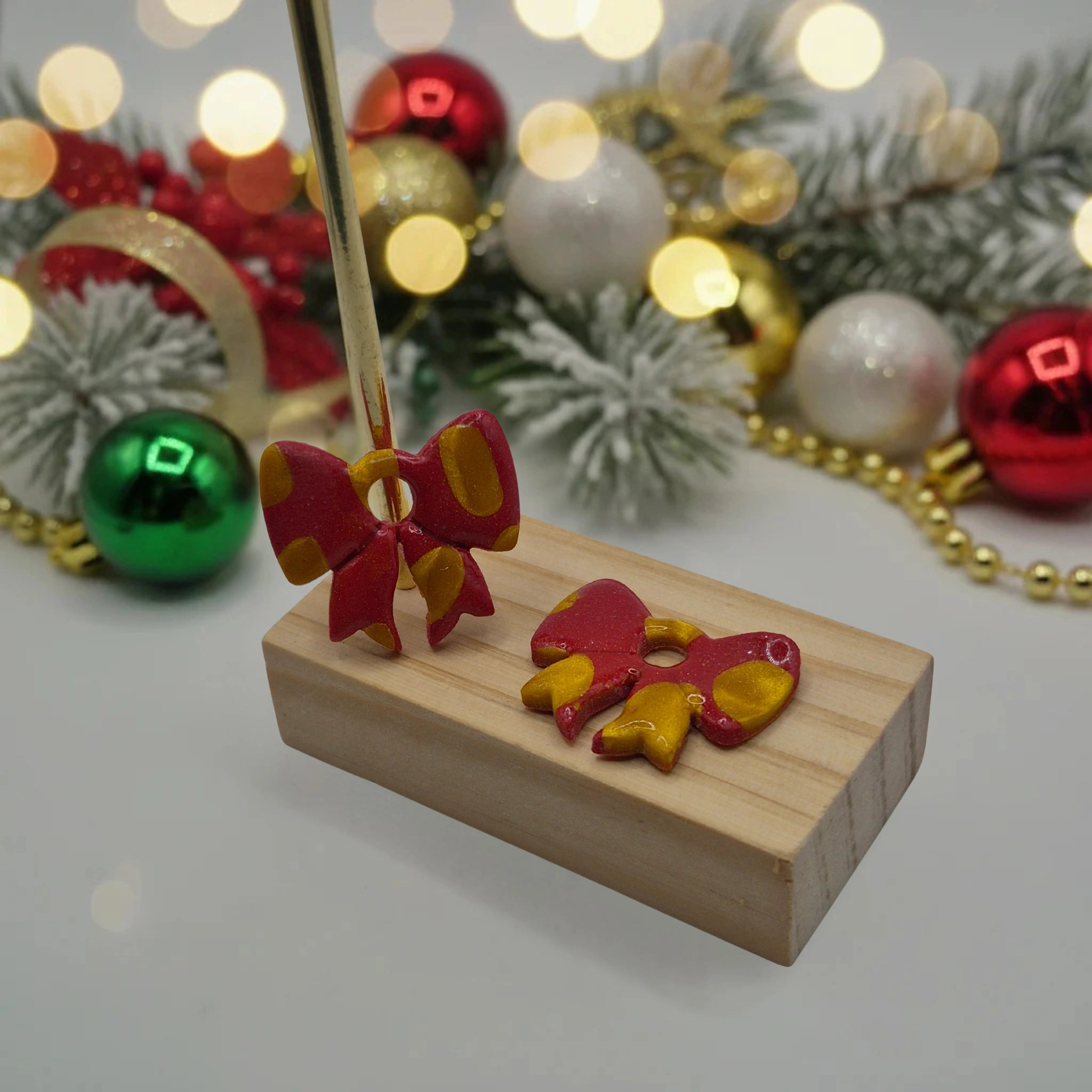 Parure Sophie : pampilles interchangeables en pâte polymère rouge et dorée pailletée, en forme de nœud, présentées sur un support en bois, et en arrière-plan des branches de sapin, des guirlandes et des boules de Noël.