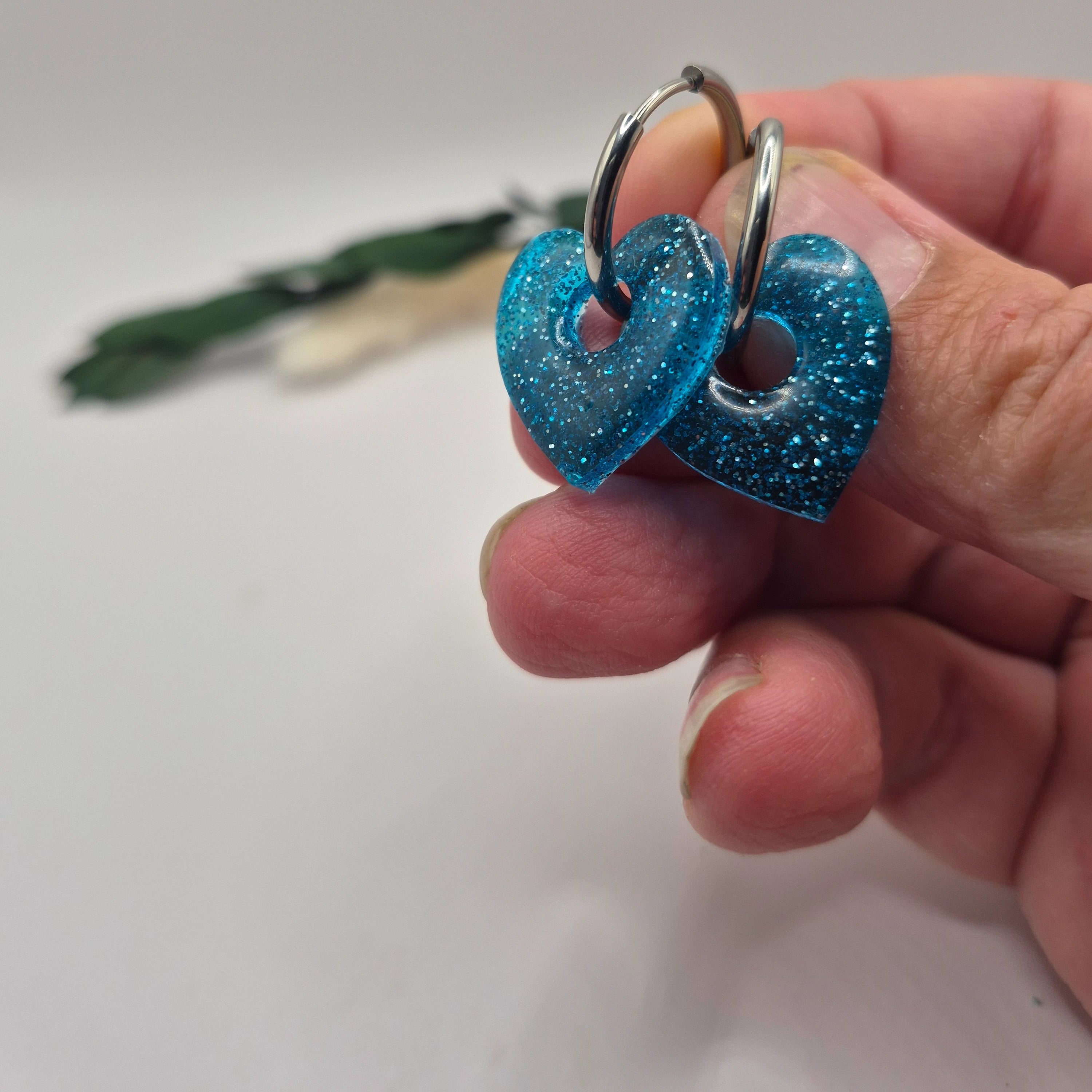 Pampilles Coralie bleues résine pailleté sur créoles inoxydables  dorées tenues à la main avec branche verte et pampa en fond de décor