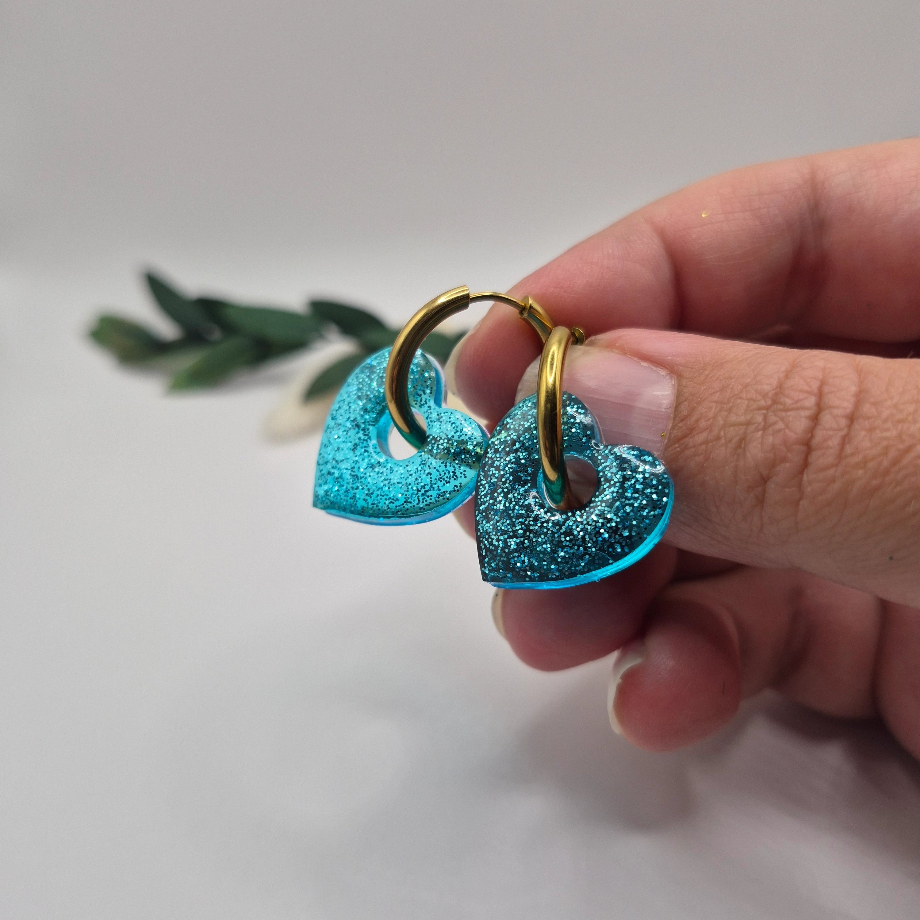 Pampilles Coralie bleues résine pailleté sur créoles inoxydables  dorées tenues à la main avec branche verte et pampa en fond de décor