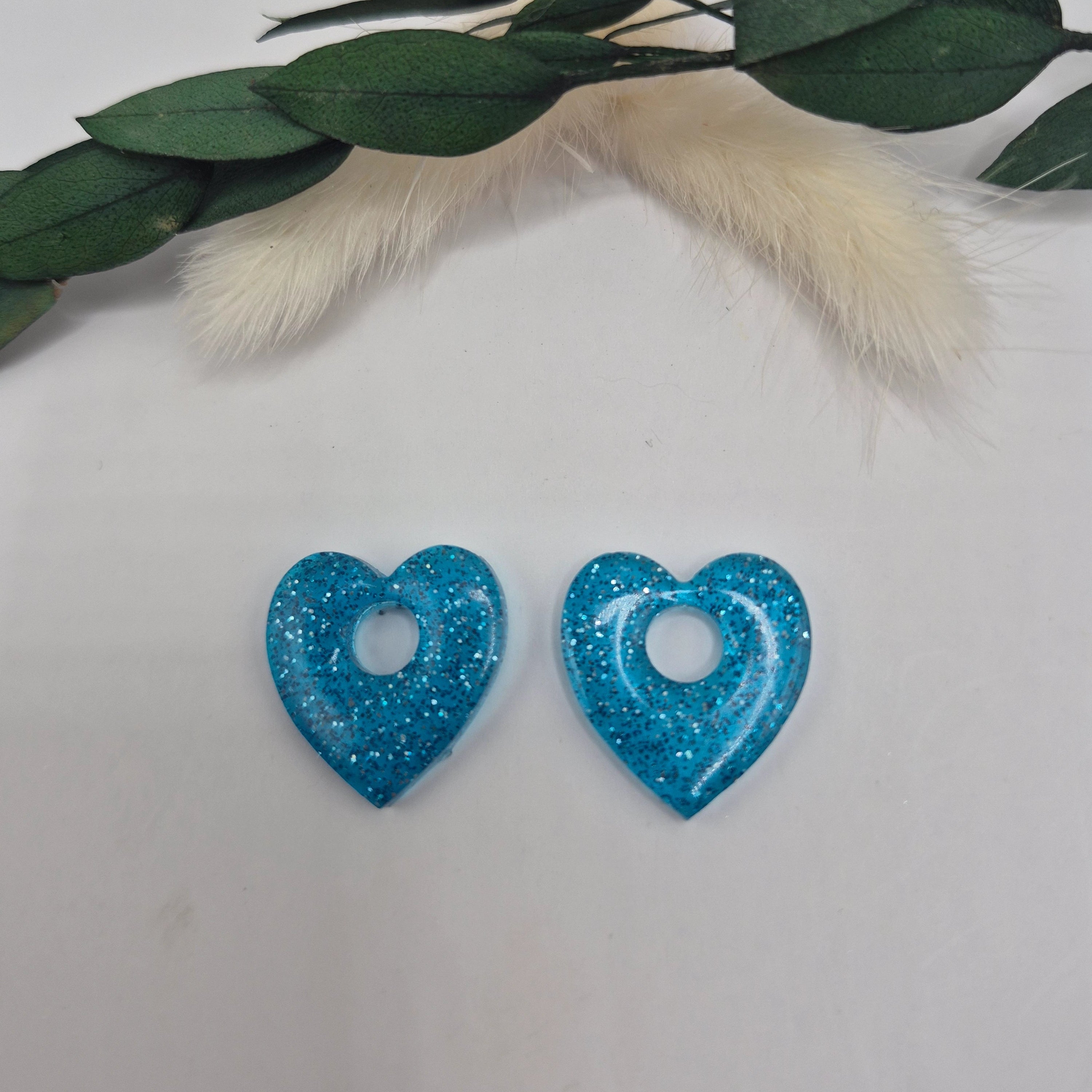 Boucles d’oreilles pampilles bleues Coralie en forme de coeur en résine époxy pailletée posées à plat sur fond neutre avec branche de pampa et feuilles