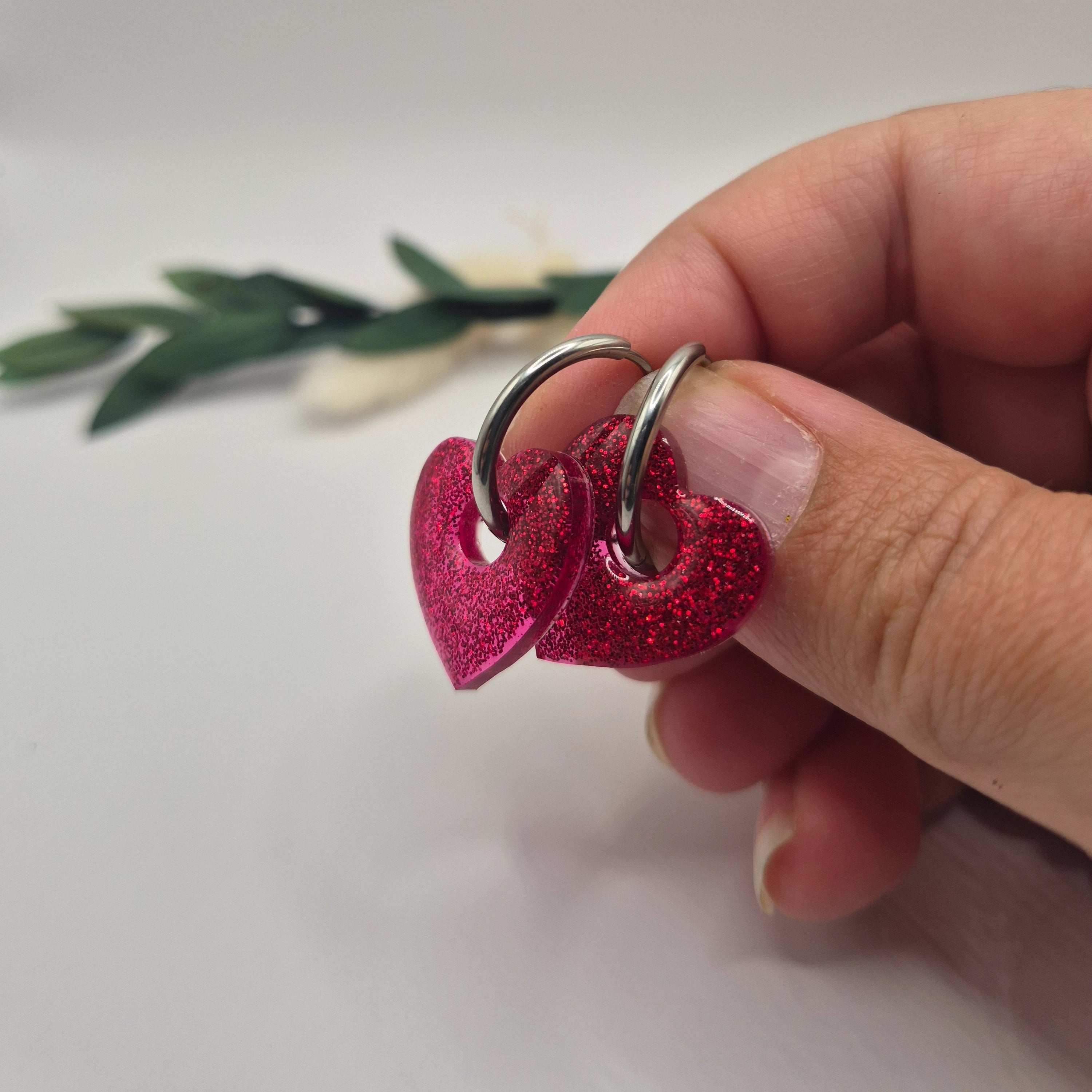 Pampilles Coralie rouges résine pailleté sur créoles inoxydables  argentées tenues à la main avec branche verte et pampa en fond de décor