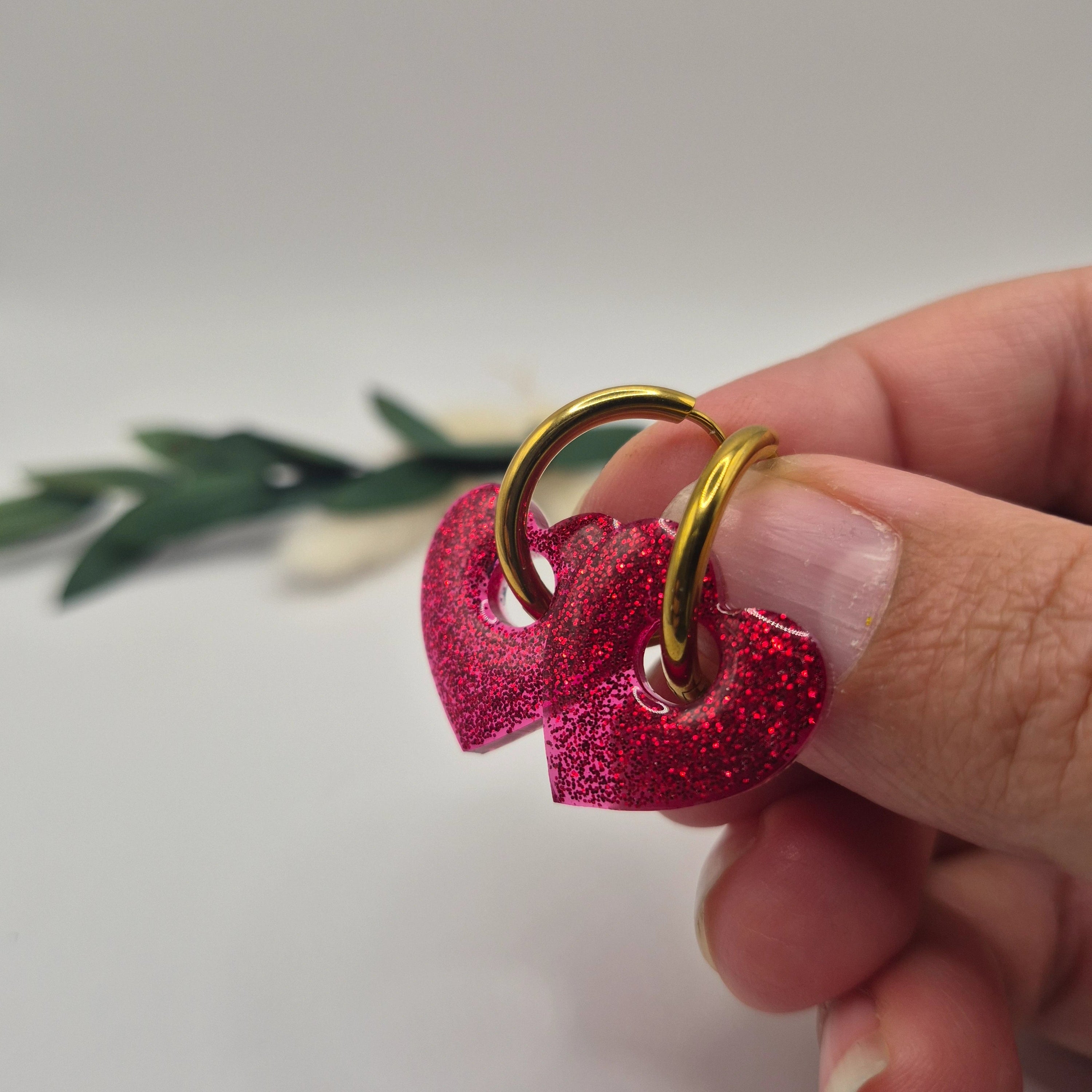 Pampilles Coralie rouges résine pailleté sur créoles inoxydables  dorées tenues à la main avec branche verte et pampa en fond de décor