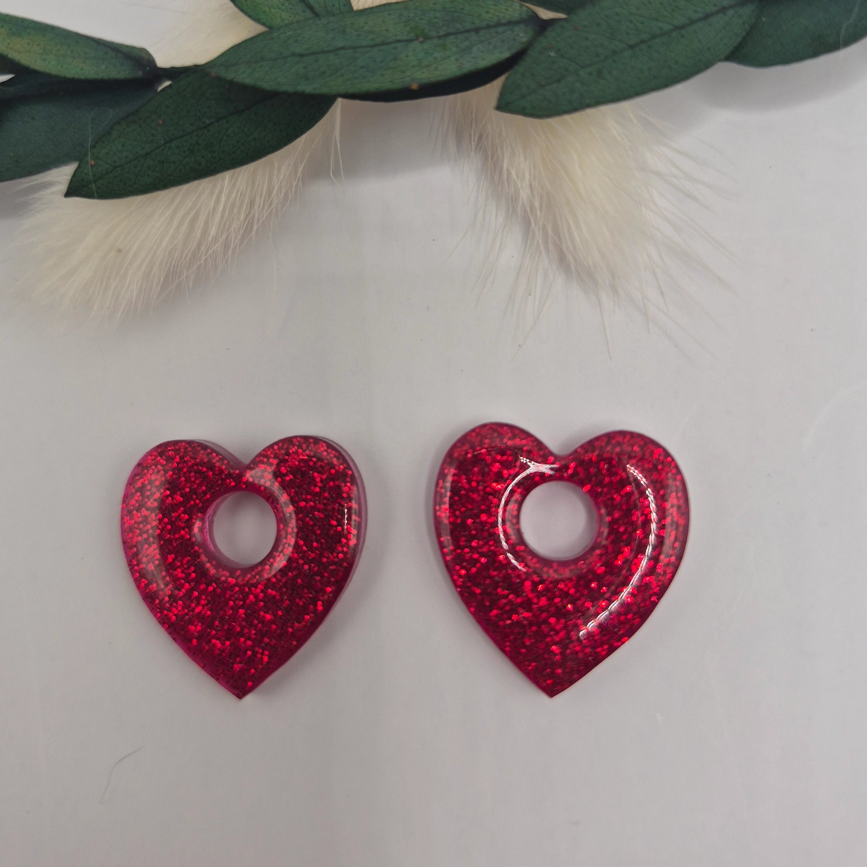 Boucles d’oreilles pampilles rouges Coralie en forme de coeur en résine époxy pailletée posées à plat sur fond neutre avec branche de pampa et feuilles