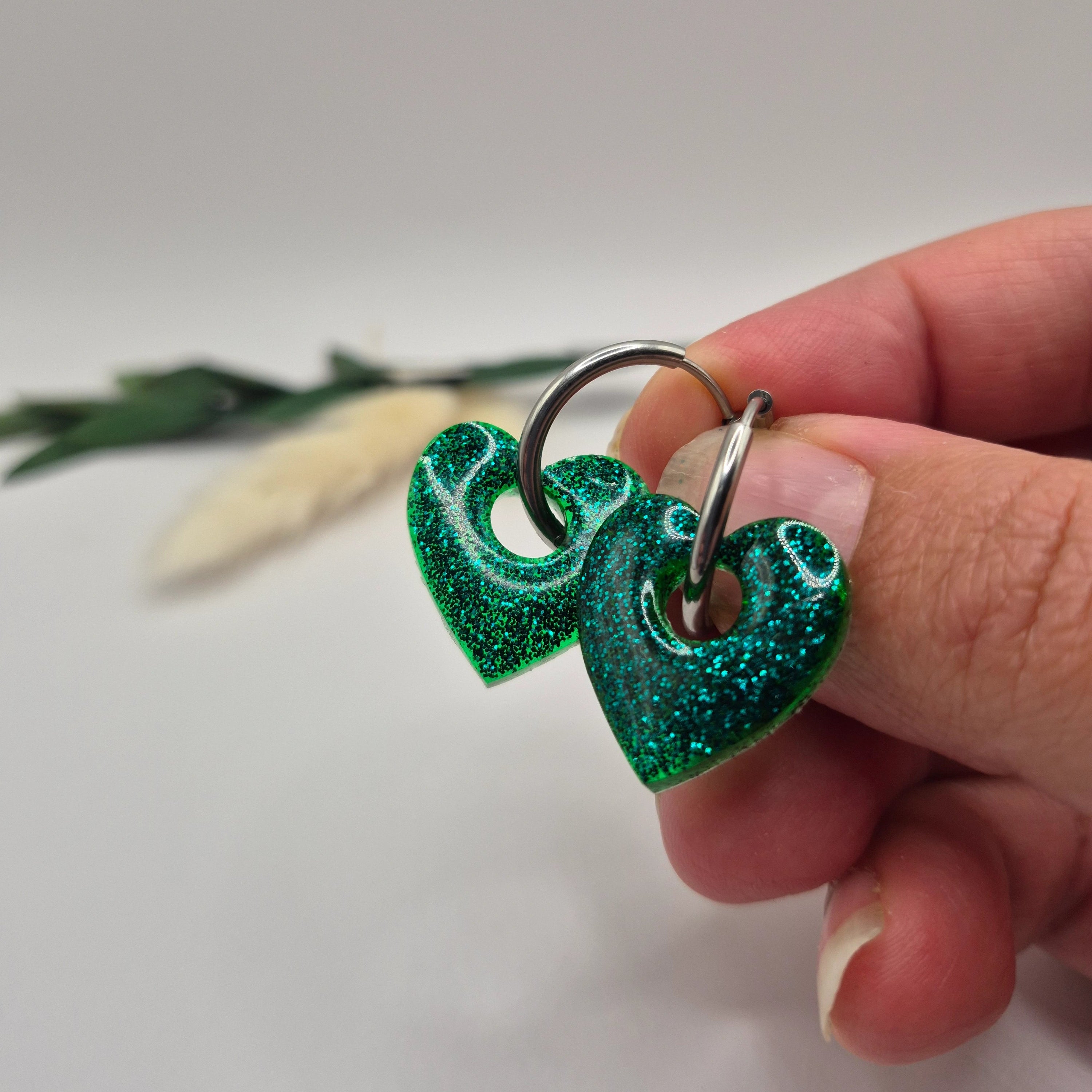 Pampilles Coralie vertes résine pailletée sur créoles inoxydables  argentées tenues à la main avec branche verte et pampa en fond de décor