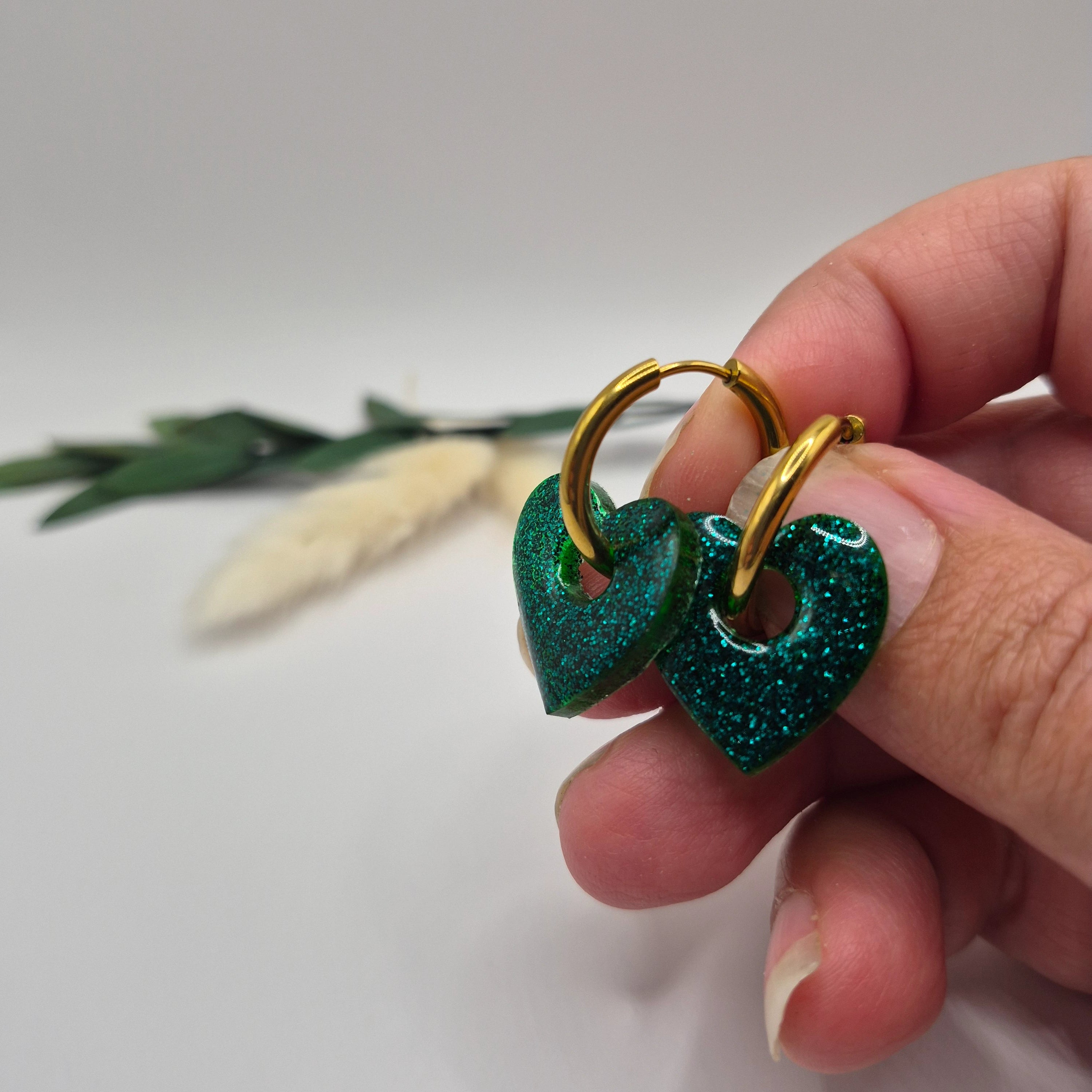 Pampilles Coralie vertes résine pailletée sur créoles inoxydables  dorées tenues à la main avec branche verte et pampa en fond de décor