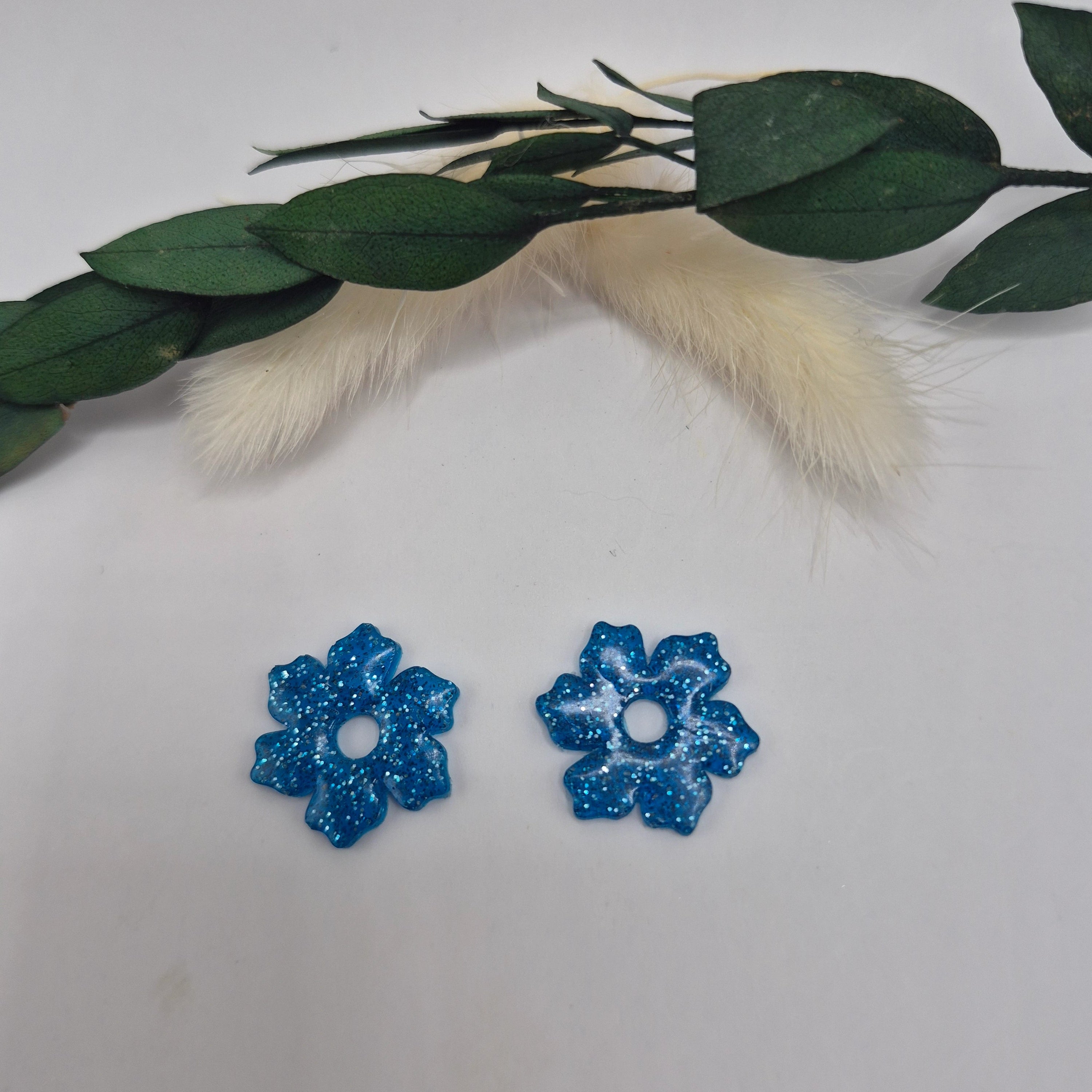 Boucles d’oreilles pampilles bleues Elodie en résine époxy pailletée posées à plat sur fond neutre avec branche de pampa et feuilles