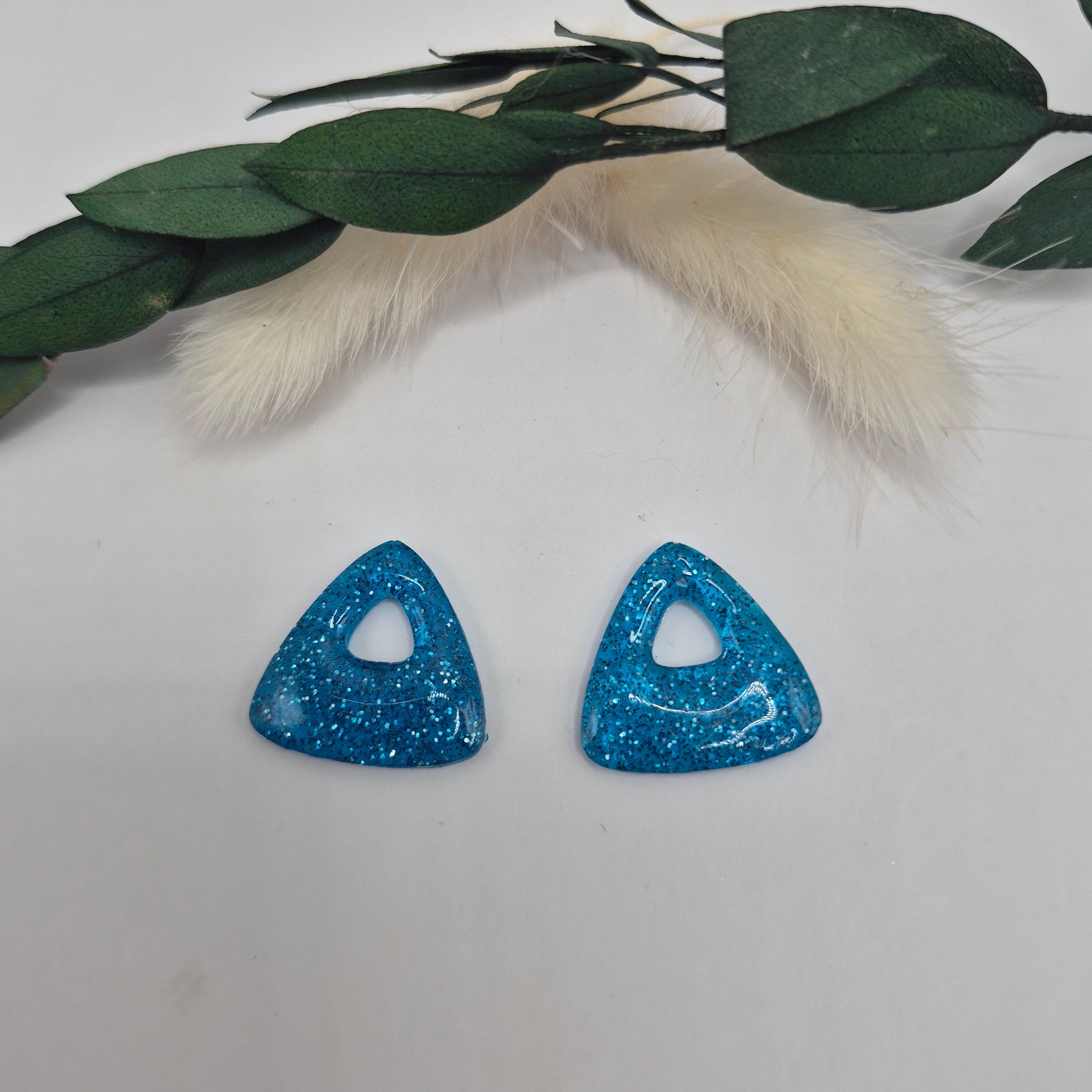Boucles d’oreilles pampilles bleues triangulaires Julie en résine époxy pailletée posées à plat sur fond neutre avec branche de pampa et feuilles