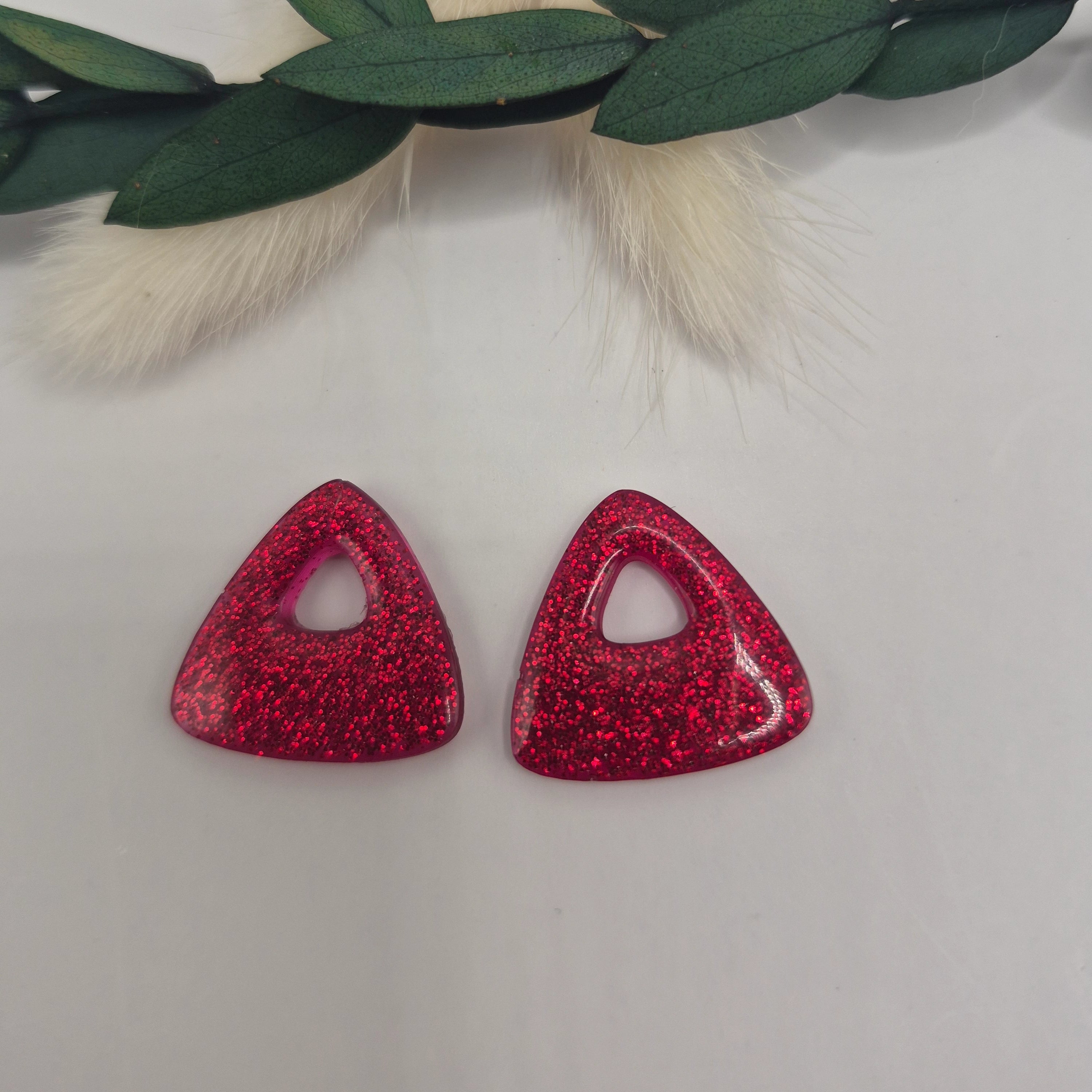 Boucles d’oreilles pampilles rouges triangulaires Julie en résine époxy pailletée posées à plat sur fond neutre avec branche de pampa et feuilles