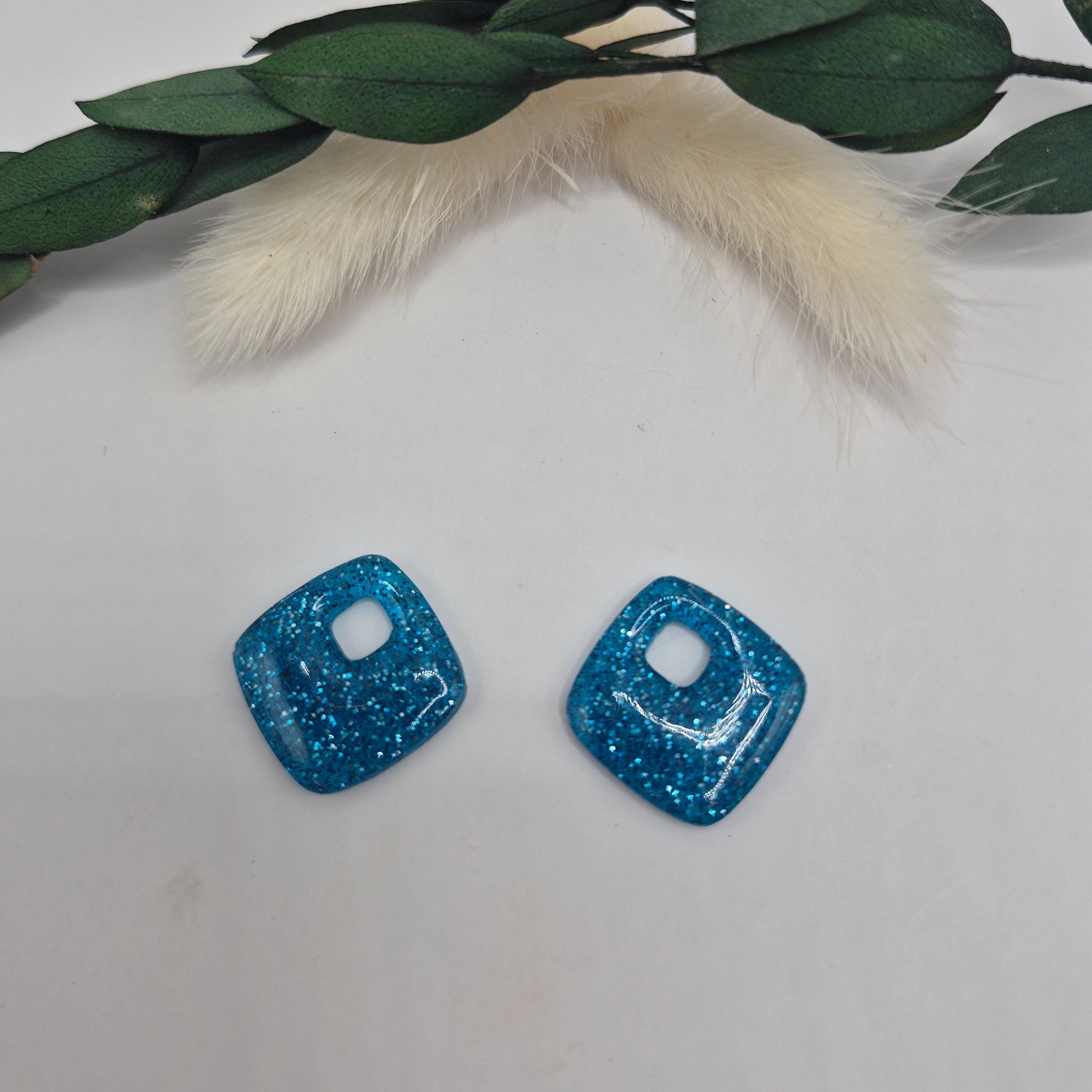Boucles d’oreilles pampilles bleues Millie en résine époxy pailletée posées à plat sur fond neutre avec branche de pampa et feuilles