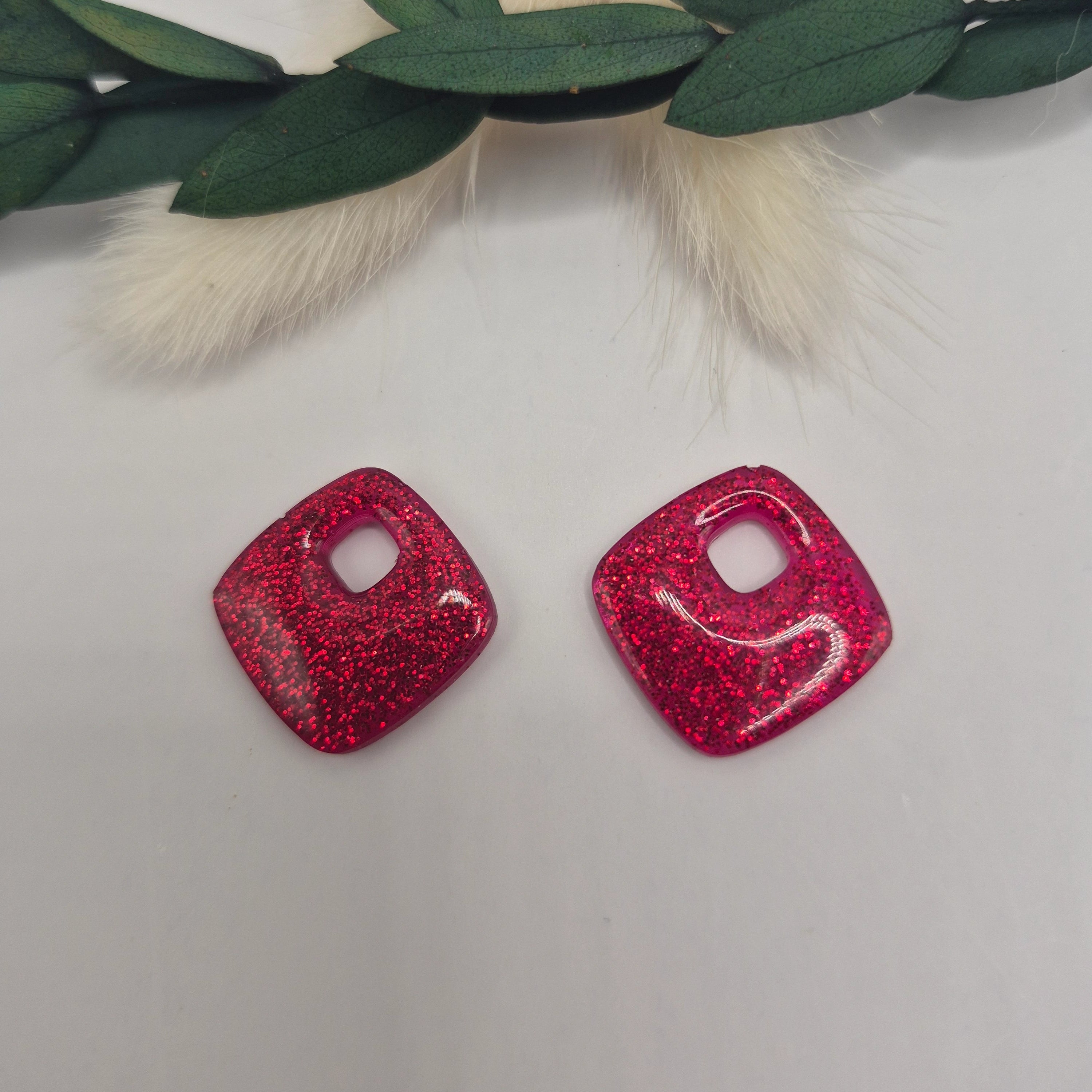 Boucles d’oreilles pampilles rouge Millie en résine époxy pailletée posées à plat sur fond neutre avec branche de pampa et feuilles