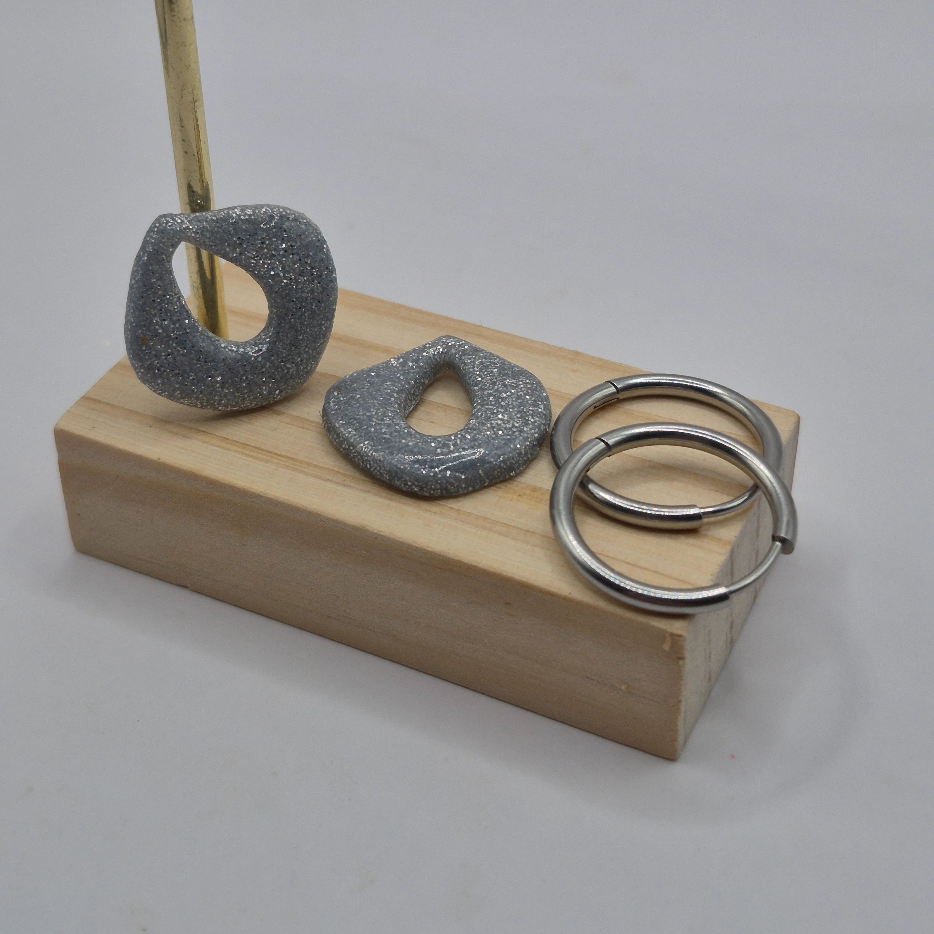 Parure Cynthia : pampilles interchangeables en pâte polymère gris argenté pailletée, en forme de goutte d'eau, présentées sur un support en bois avec deux créoles inoxydables argentées.