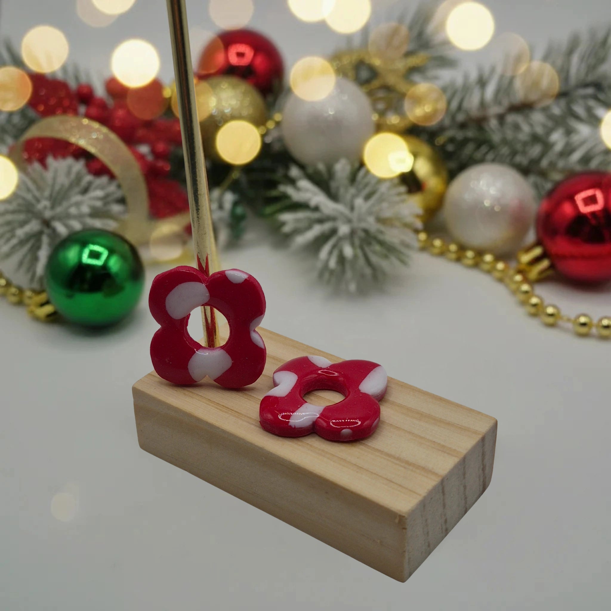 Parure Carine : pampilles interchangeables en pâte polymère rouge et blanc, en forme de fleur, présentées sur un support en bois avec en arrière-plan des branches de sapin, des guirlandes et des boules de Noël.