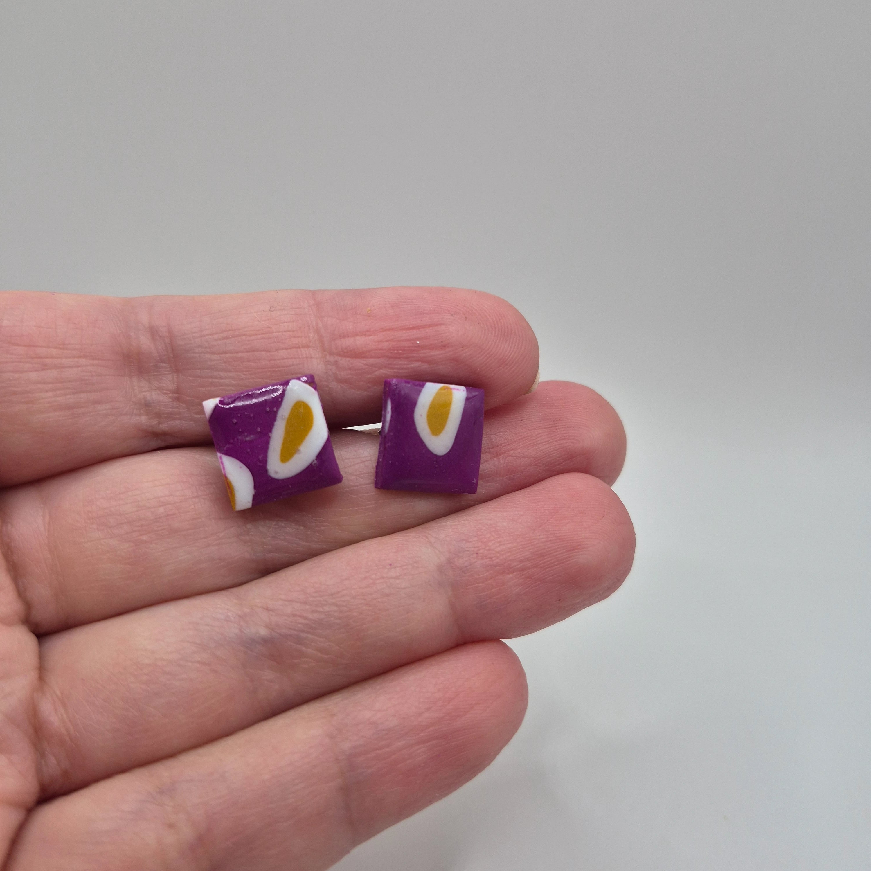 Boucles d’oreilles avec petites pampilles carrées en pâte polymère violette, faites main et finition résine, présentées dans la main — parure artisanale Emma.