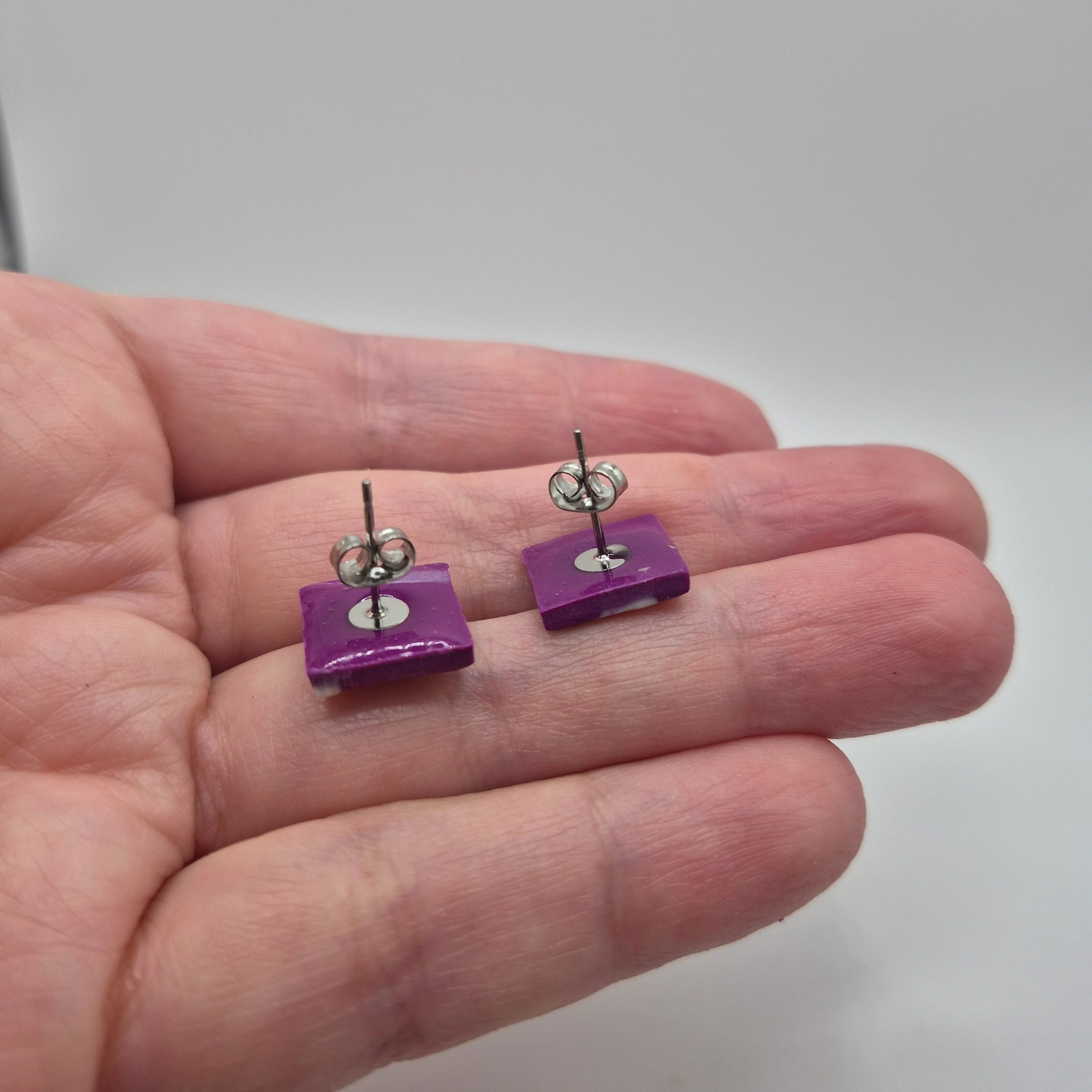 Boucles d’oreilles artisanales en pâte polymère violette, petites pampilles carrées finition résine, présentées dans la main avec un zoom sur l’attache à clou en acier inoxydable.