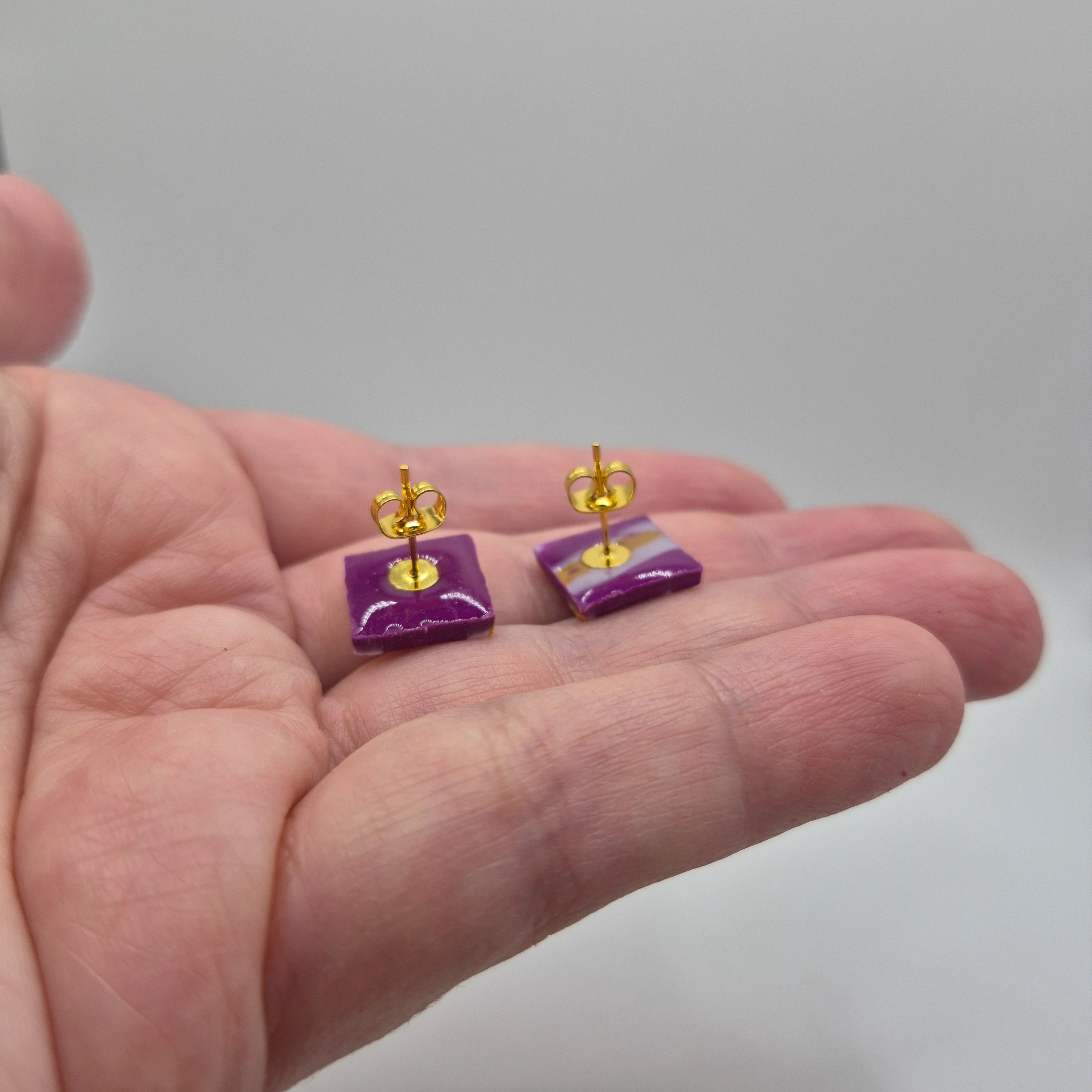 Boucles d’oreilles artisanales en pâte polymère violette, petites pampilles carrées finition résine, présentées dans la main avec un zoom sur l’attache dorée à clou en acier inoxydable.
