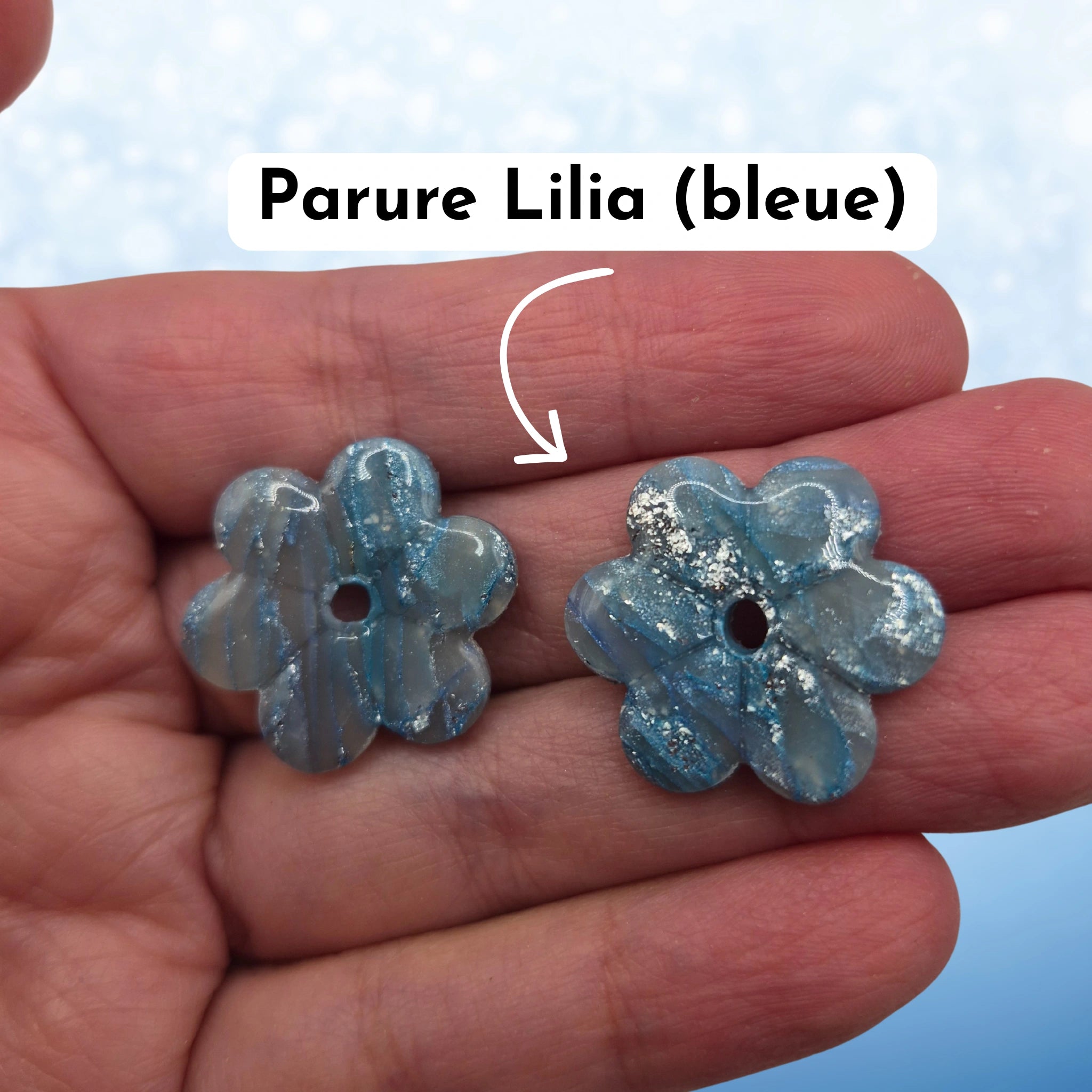 Pampilles en pâte polymère bleues, en forme de fleur, avec l’inscription « Parure Lilia (bleue) », présentées tenues dans une main, sur un fond en dégradé de bleu.