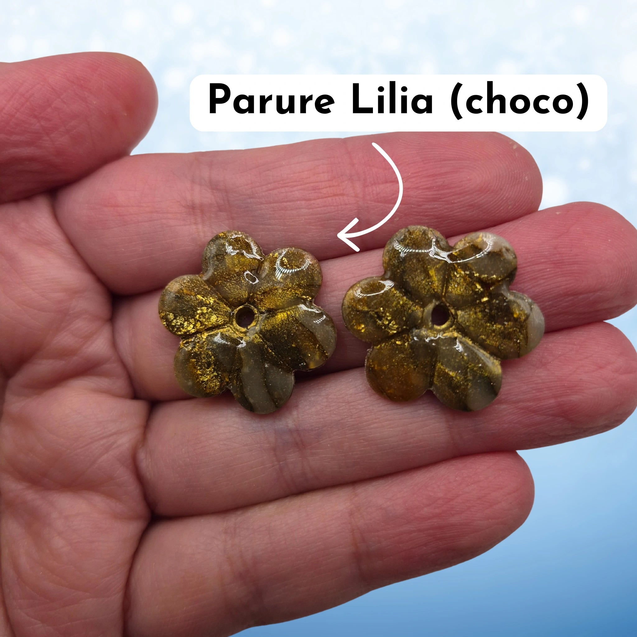 Pampilles en pâte polymère marrons, en forme de fleur, avec l’inscription « Parure Lilia (choco) », présentées tenues dans une main, sur un fond en dégradé de bleu.