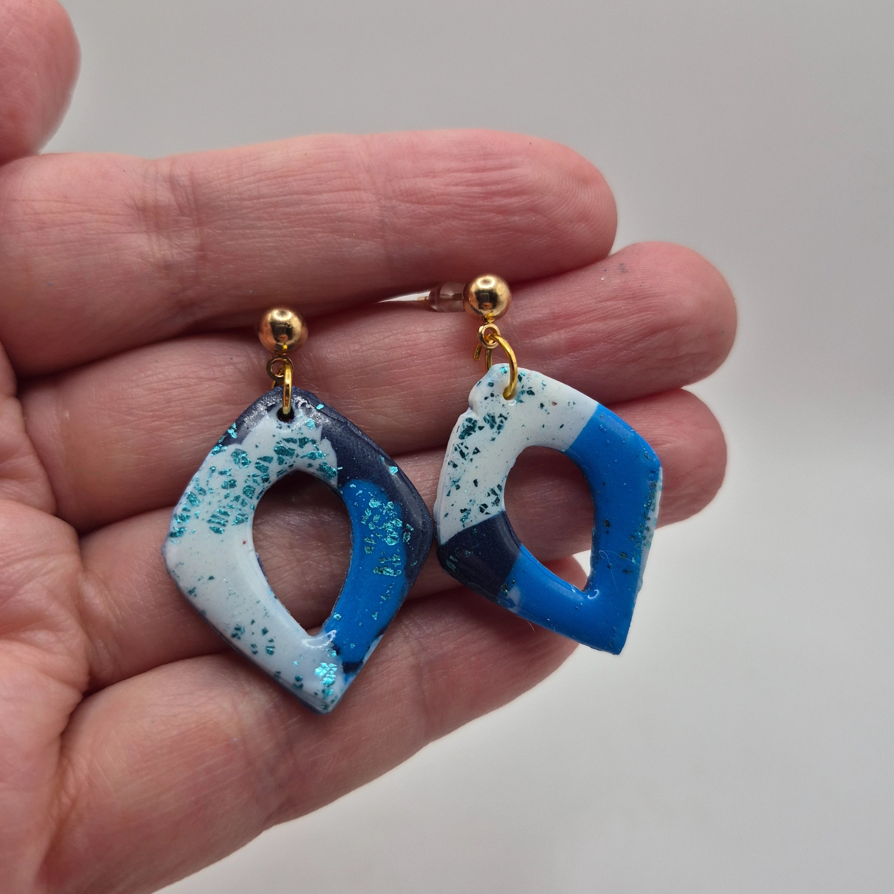 Boucle d'oreilles ou Pampilles Marcia en pâte polymère bleue et blanche, finition résine, de forme losange, avec attaches à clous en acier inoxydable doré, présentées à la main.
