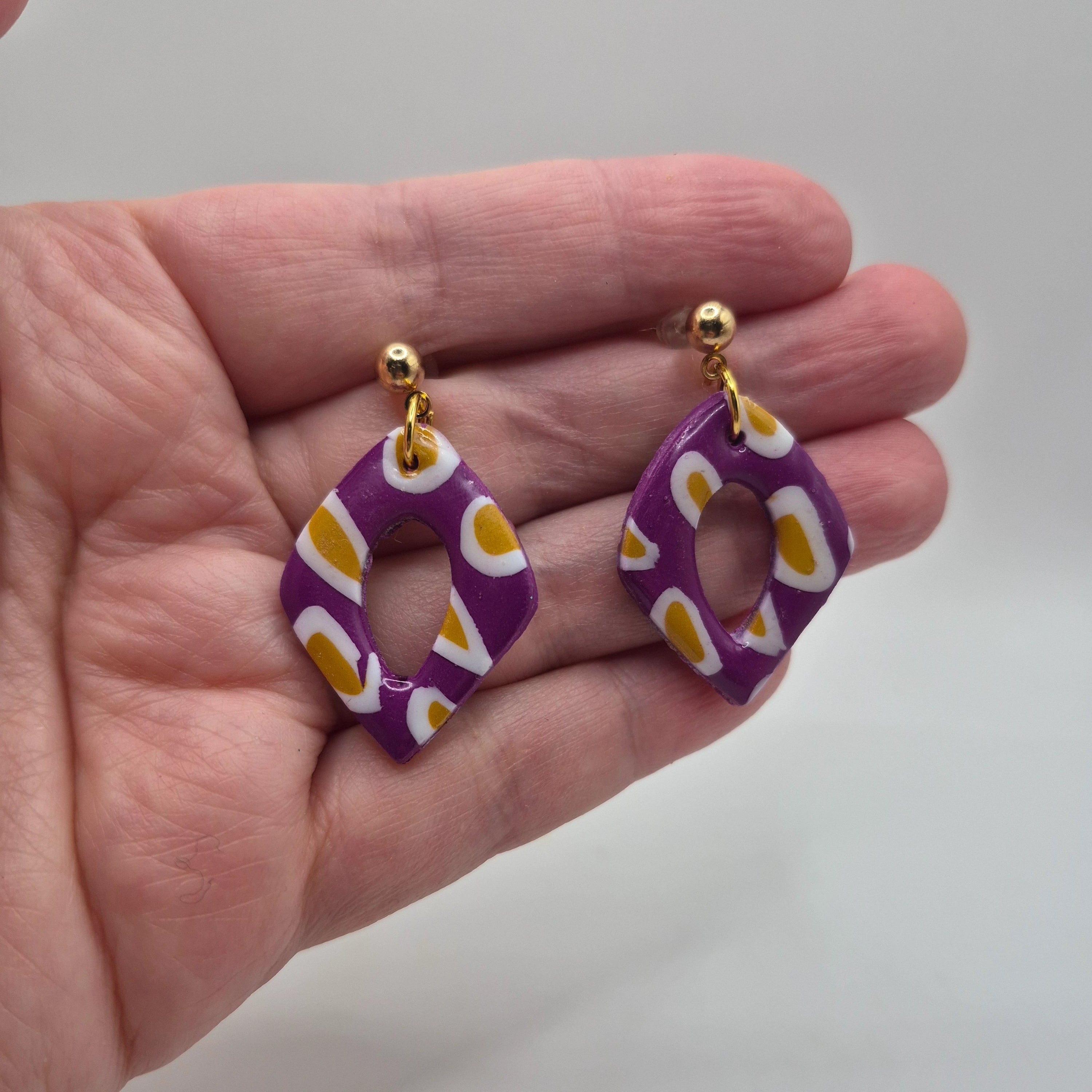 Boucle d'oreilles ou Pampilles Marcia en pâte polymère violette avec motif ocre et blanc, finition résine, de forme losange, avec attaches à clous en acier inoxydable doré, présentées à la main.