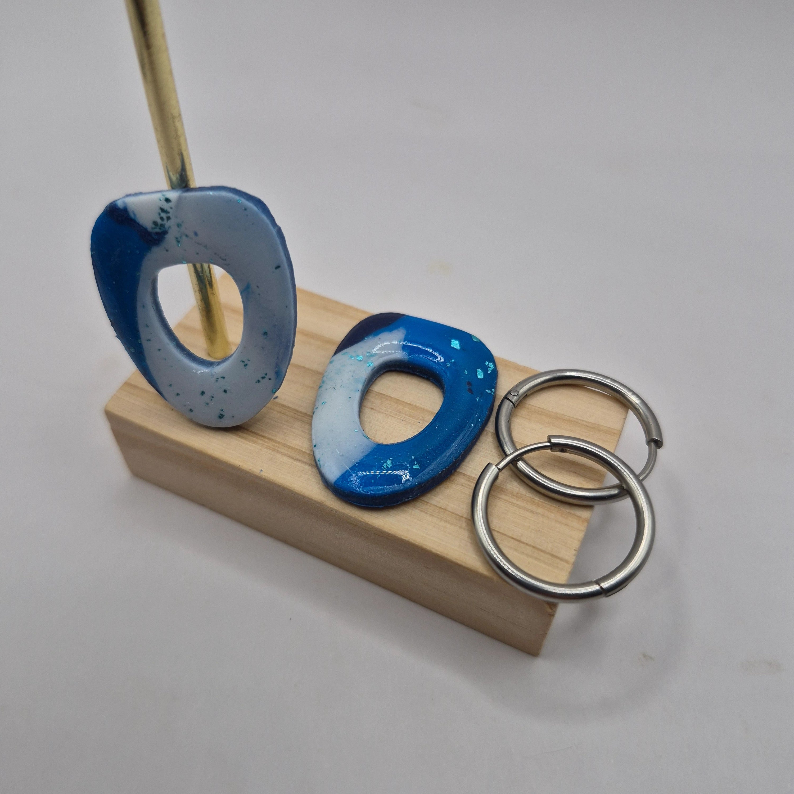 Boucles d’oreilles pampilles en pâte polymère bleu et blanc, de forme arrondie et géométrique, présentées sur un support en bois à côté de deux créoles en acier inoxydable argenté — parure Mia.