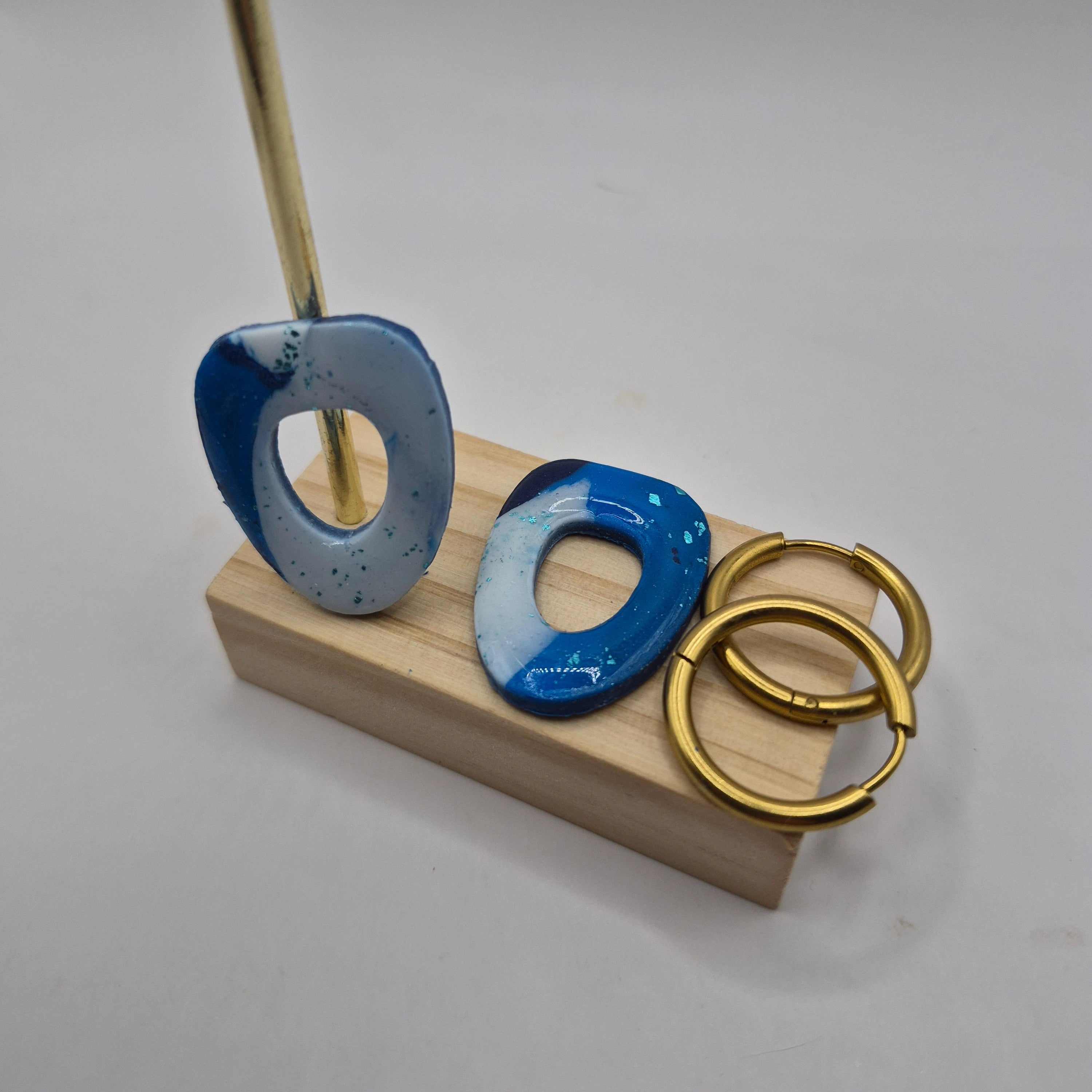 Boucles d’oreilles pampilles en pâte polymère bleu et blanc, de forme arrondie et géométrique, présentées sur un support en bois à côté de deux créoles en acier inoxydable doré — parure Mia.