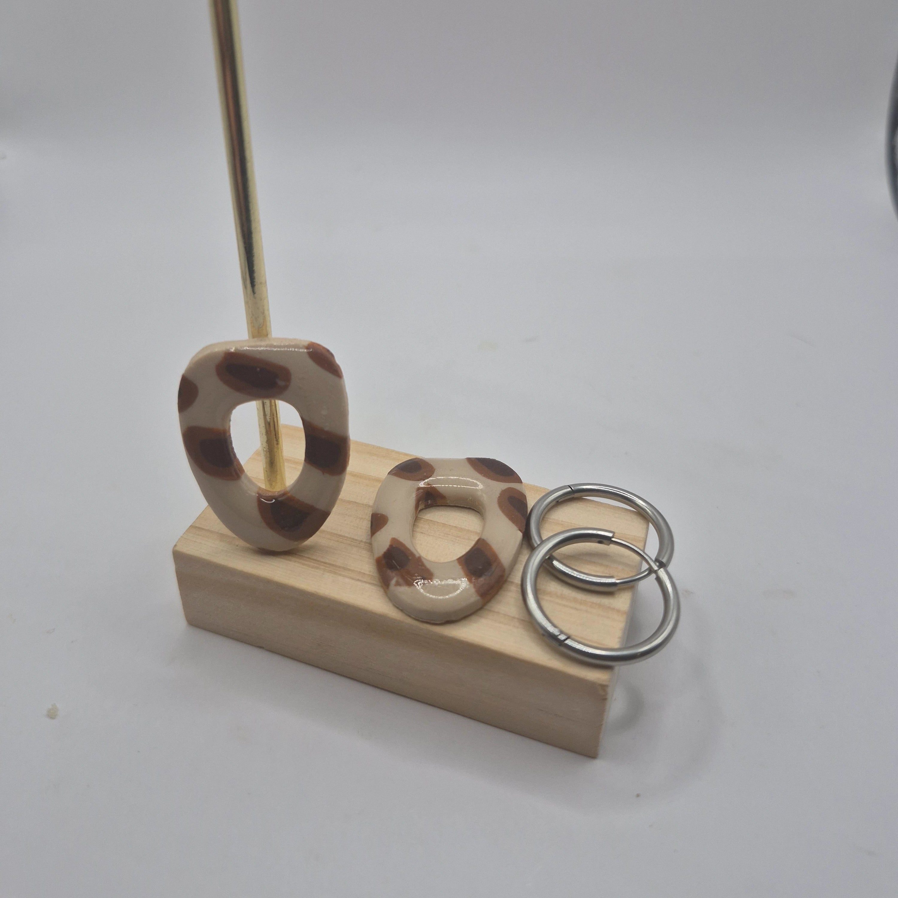 Boucles d’oreilles pampilles en pâte polymère motif léopard , de forme arrondie et géométrique, présentées sur un support en bois à côté de deux créoles en acier inoxydable argenté — parure Mia.