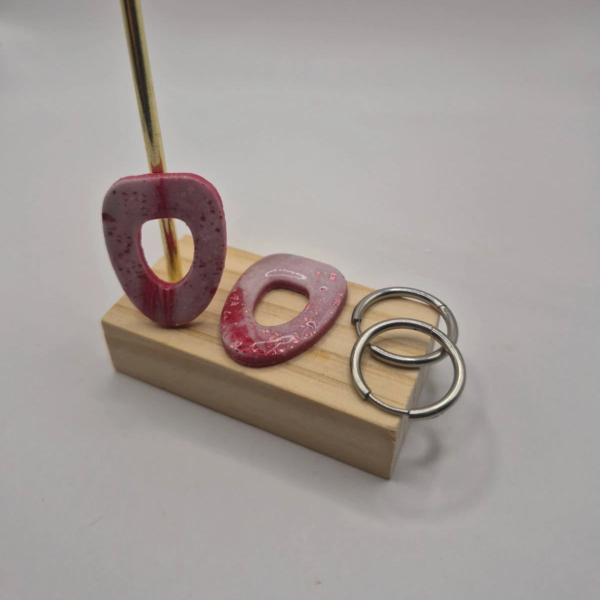 Boucles d’oreilles pampilles en pâte polymère rose, de forme arrondie et géométrique, présentées sur un support en bois à côté de deux créoles en acier inoxydable argenté — parure Mia.
