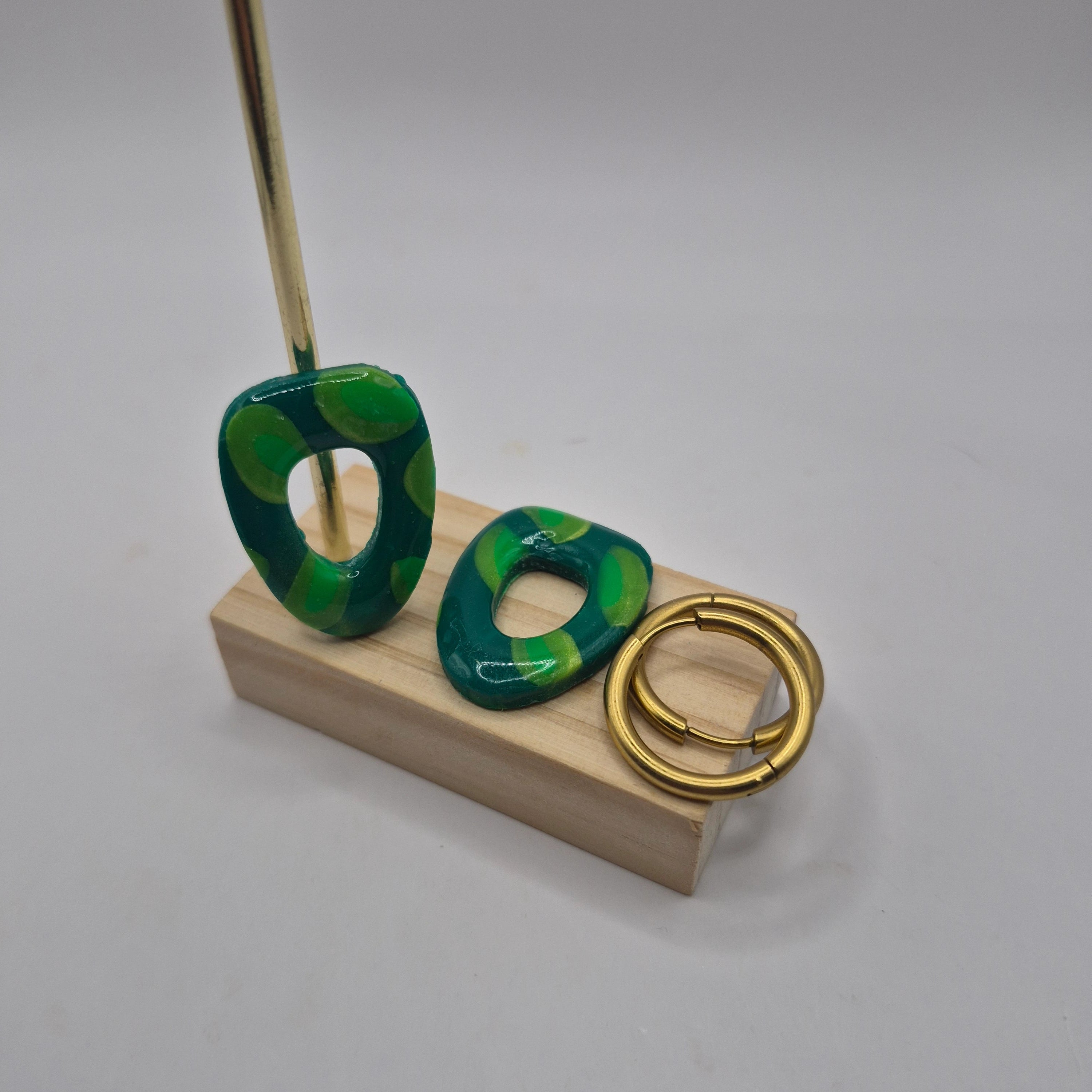 Boucles d’oreilles pampilles en pâte polymère vert, de forme arrondie et géométrique, présentées sur un support en bois à côté de deux créoles en acier inoxydable doré — parure Mia.