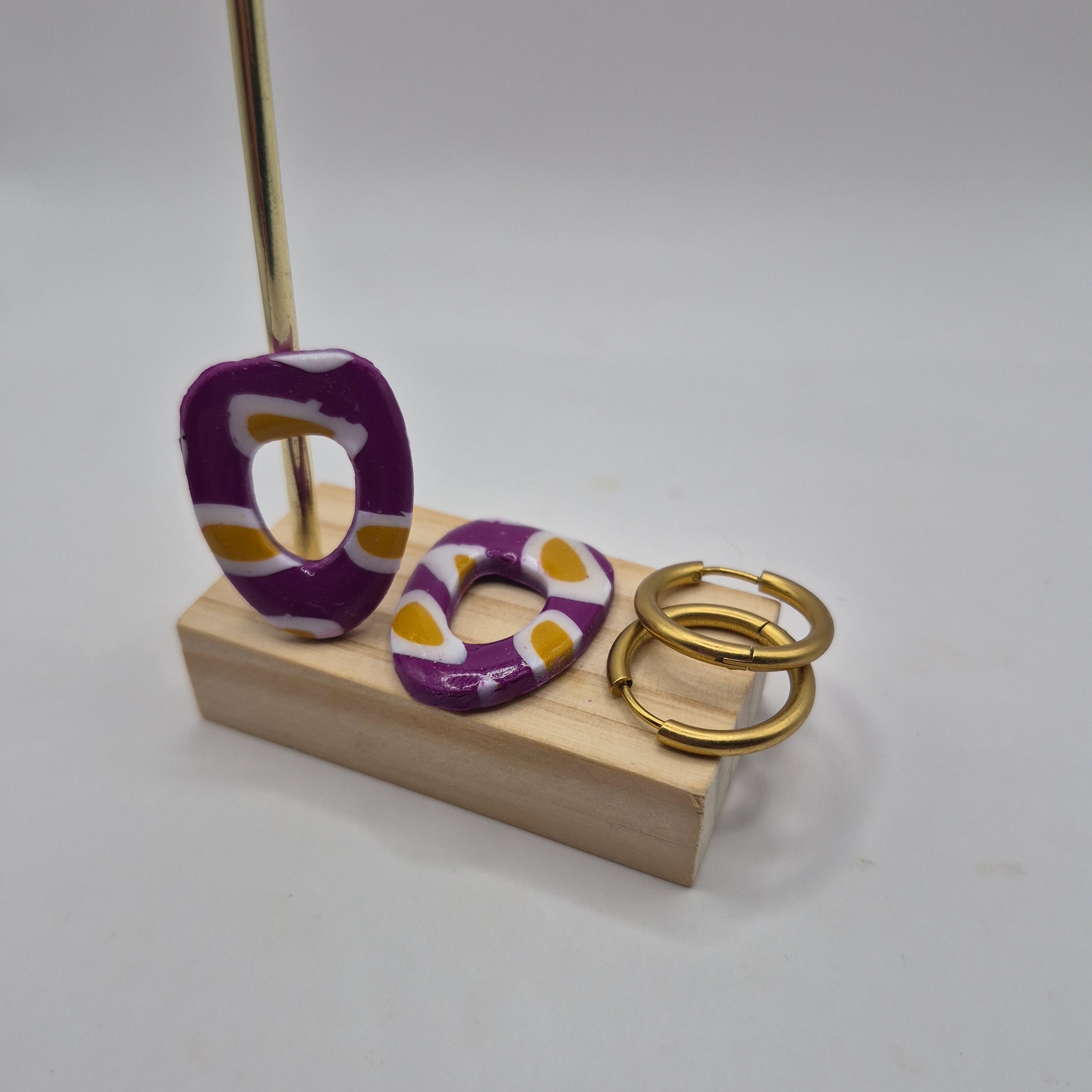 Boucles d’oreilles pampilles en pâte polymère violet, de forme arrondie et géométrique, présentées sur un support en bois à côté de deux créoles en acier inoxydable doré — parure Mia.