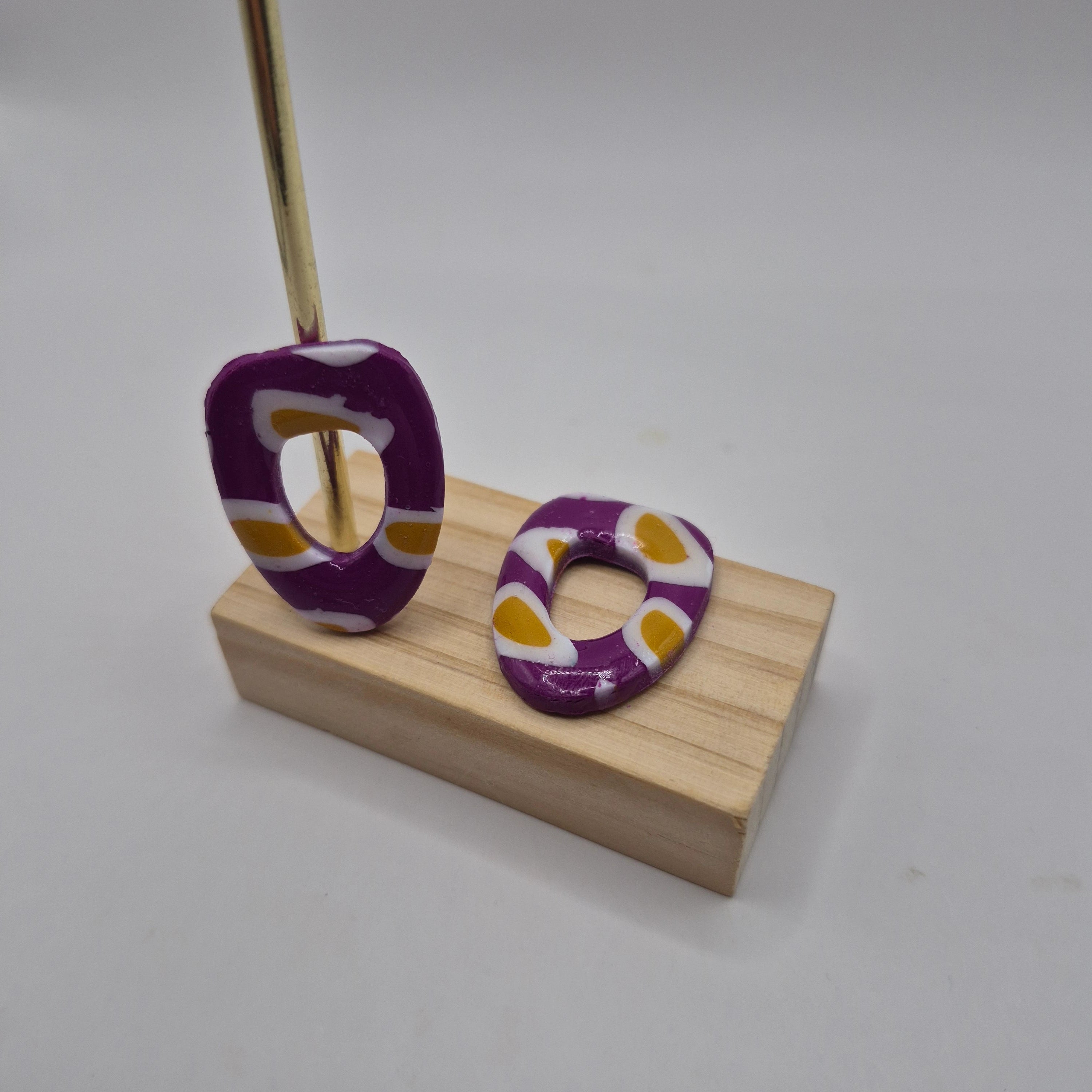 Boucles d’oreilles pampilles en pâte polymère violet, de forme arrondie et géométrique, présentées sur un support en bois — parure Mia.